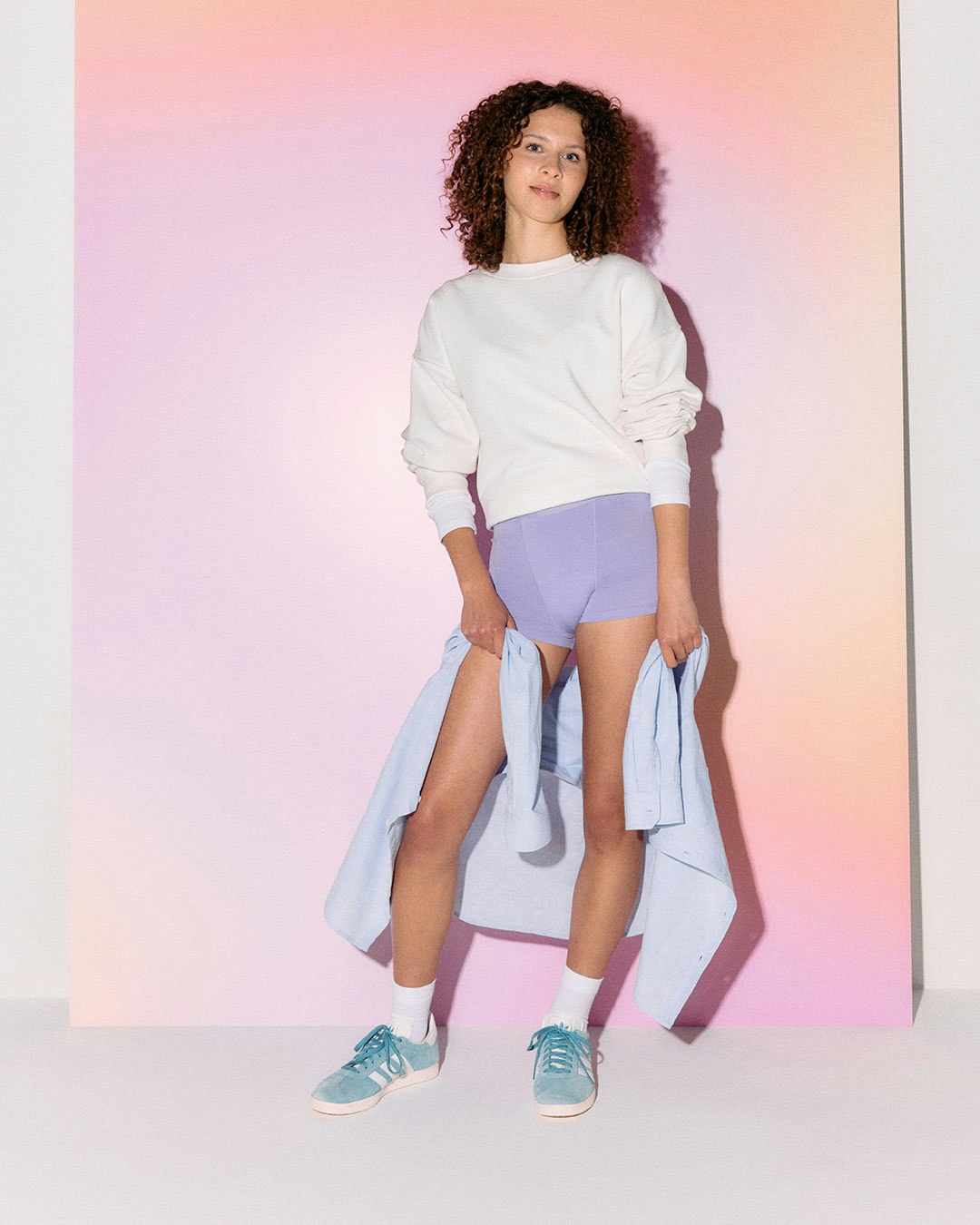 A person stands in front of a pastel gradient backdrop, wearing a white sweatshirt and purple period panties.