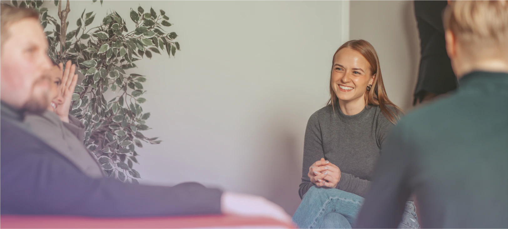 A casual office or meeting setting where a woman in a grey sweater with straight hair sits smiling warmly while engaging in conversation with colleagues, who are partially visible in the foreground. A decorative plant with green leaves is visible in the background against a light-colored wall.