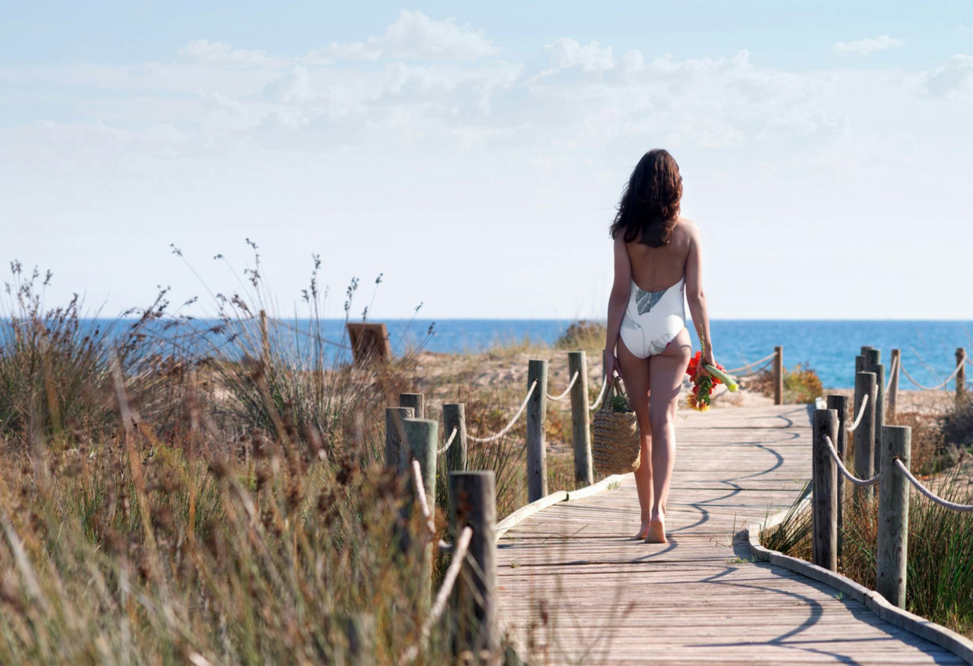 Woman walking down to the ocean wearing a white one piece bathing suit