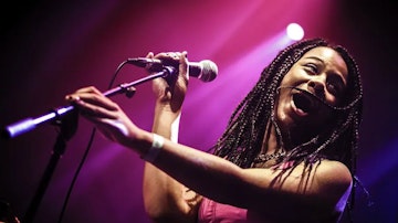 Muzikant Pop studente Adura met microfoon in haar hand op het podium met een roze paars licht achter haar.