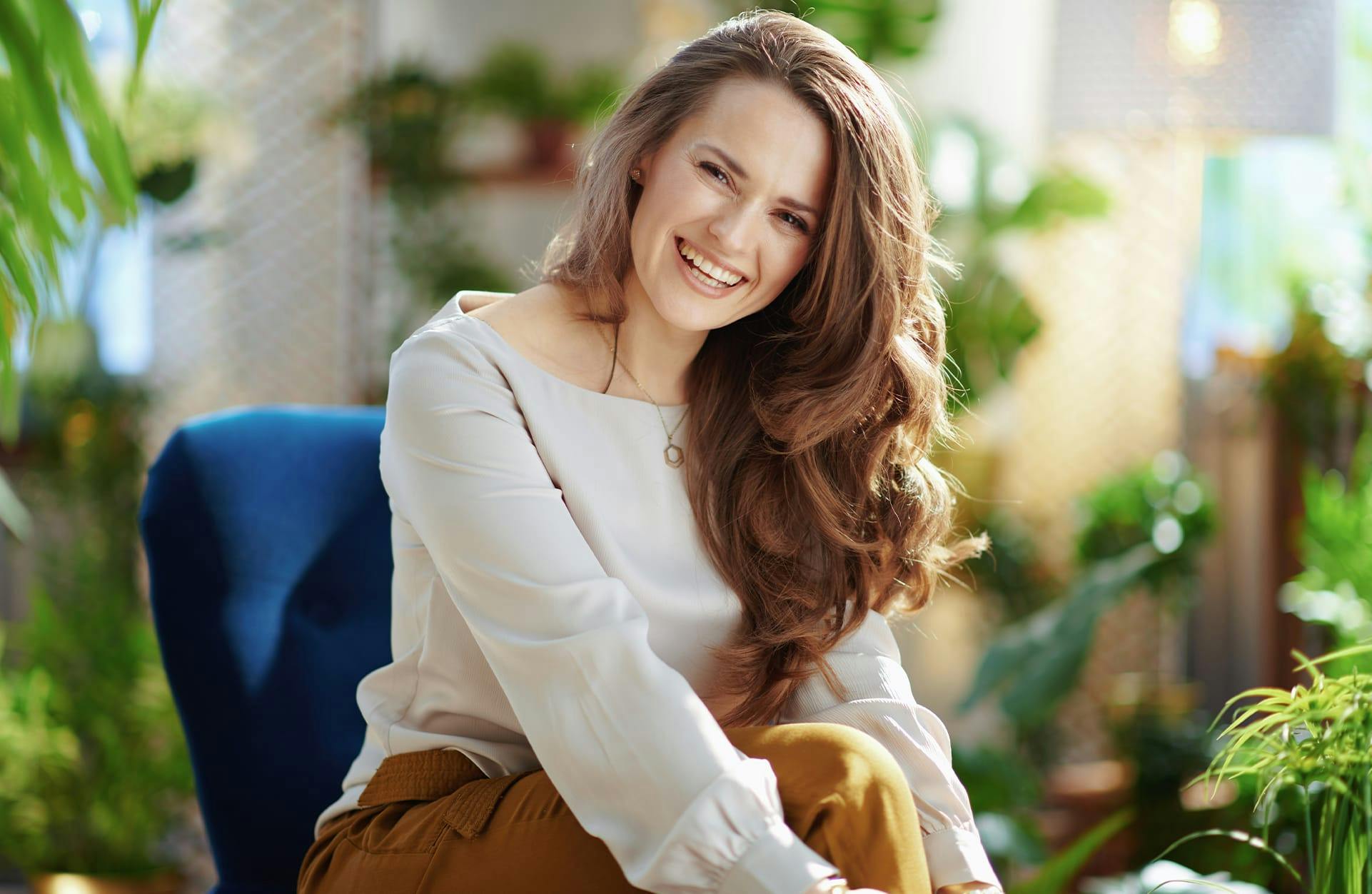 brunette woman sitting on a blue chair, tan top and brown pants laughing