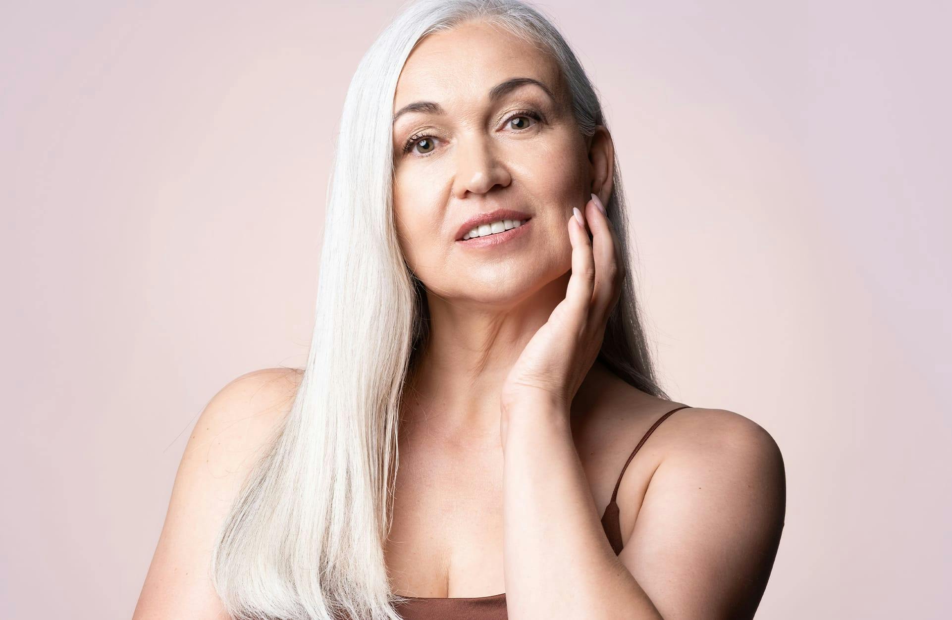 older woman with long grey hair half smiling at the camera with her hand on her face