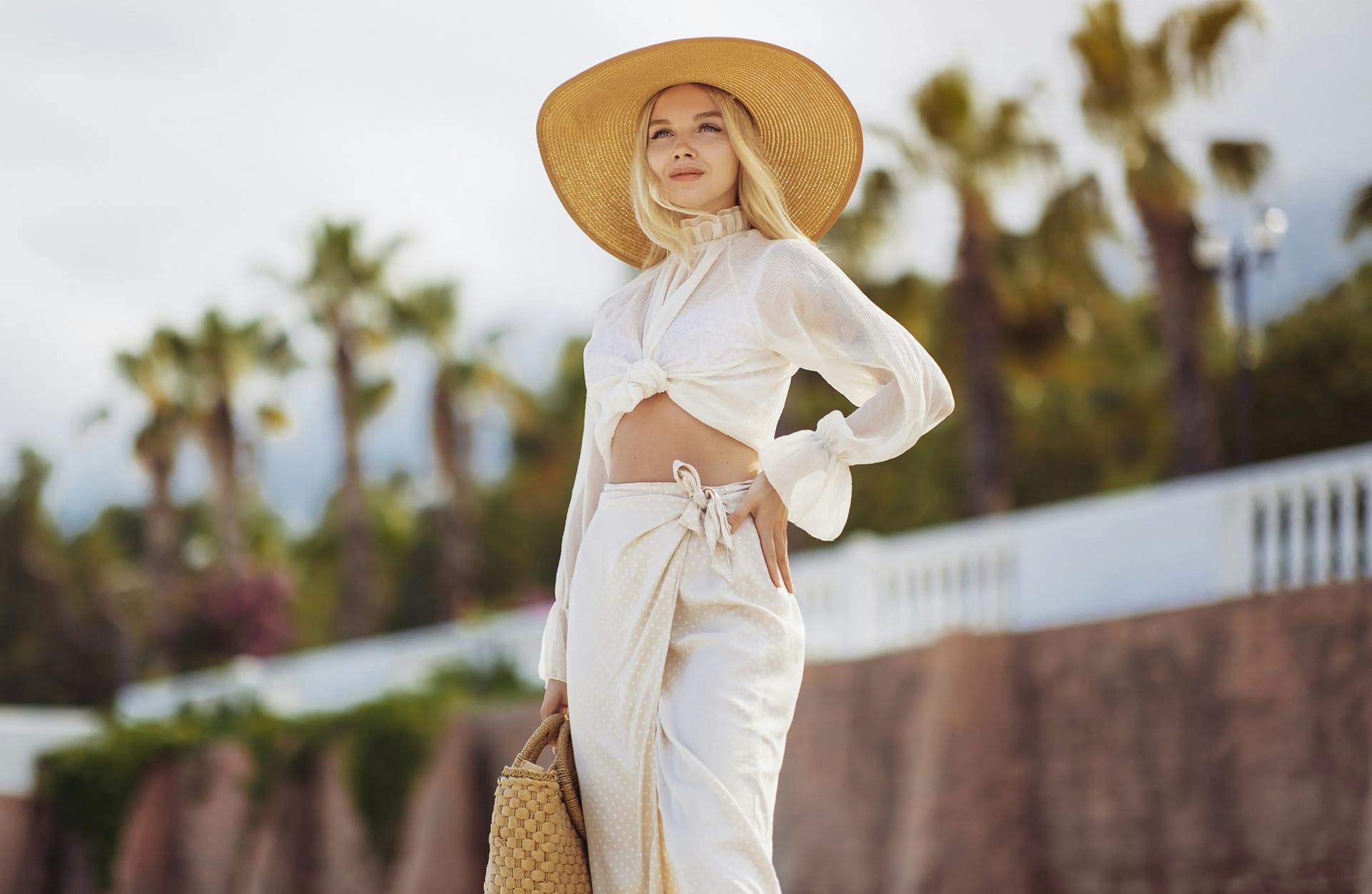 blonde woman standing in a white outfit and large hat with her hand on her hip