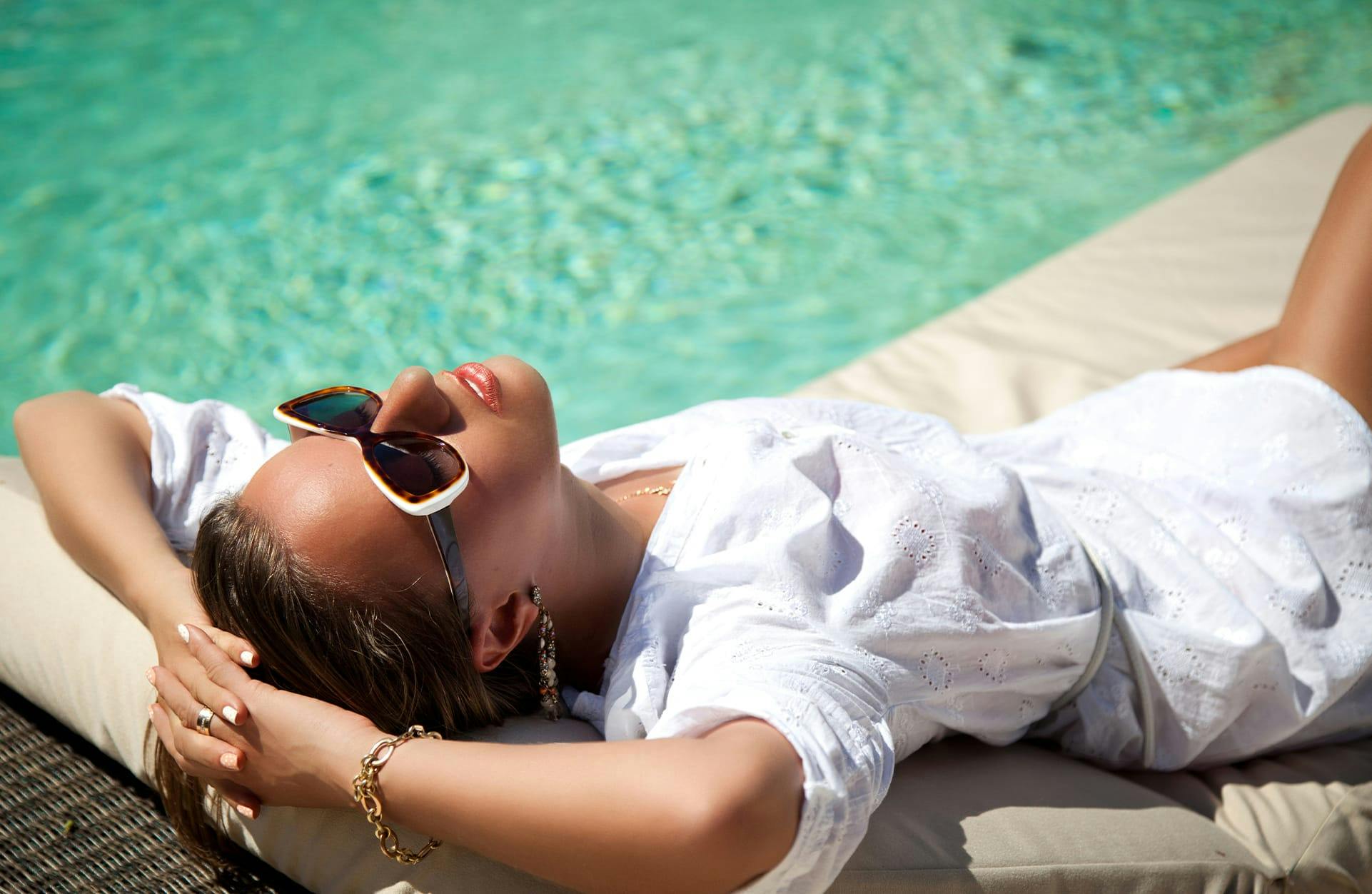 brunette woman laying outside by the pool in sunglasses and a white dress
