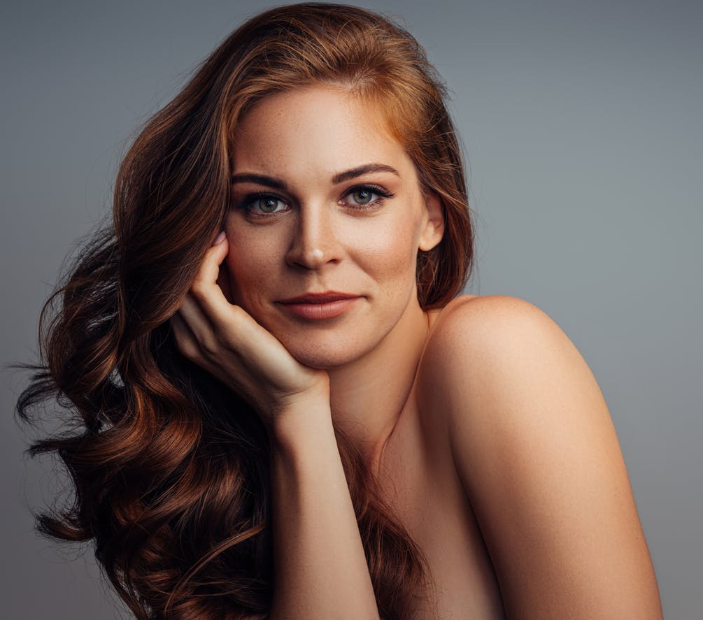 Close up of young woman's face and upper part of chest. She is smiling with one hand up on her cheek and has perfect, clear skin. Her hands rest gently around her face. Her beatiful skin is to emulate the potential results of Toskani Skincare in Austin, TX with APSI