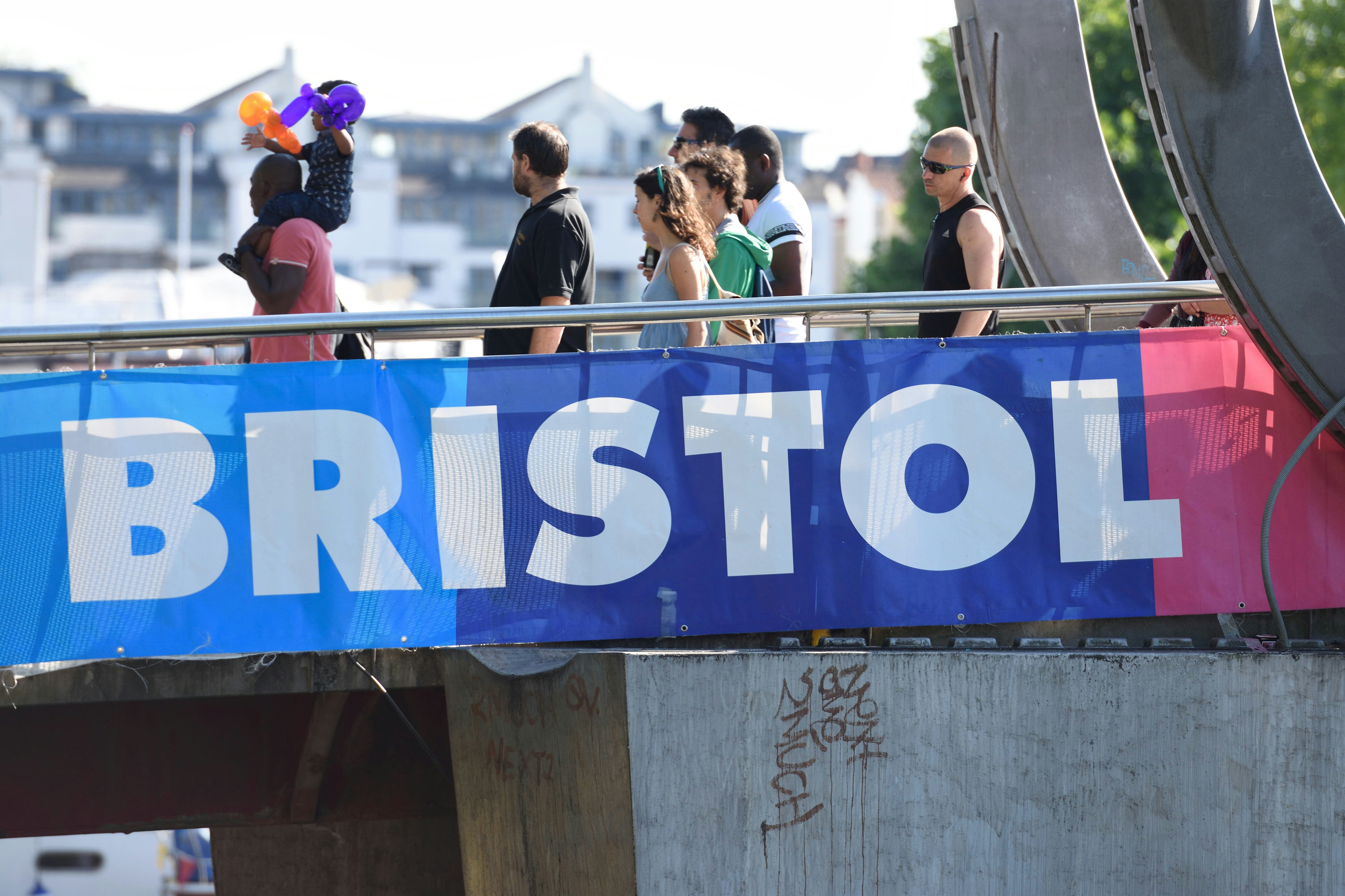 People of different ages, races and genders walking across a bridge. The bridge has a banner with the word 'Bristol' on its side.
