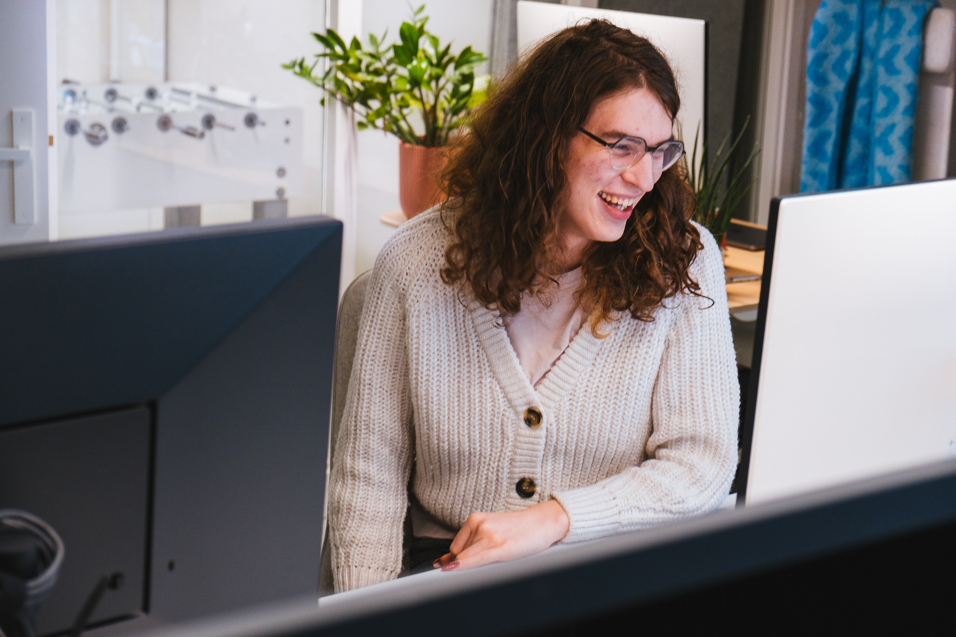 A woman with curly hair and glasses smiles while sitting at a desk, looking at her computer screen. She is wearing a cozy white cardigan and appears to be in a modern office setting with a potted plant and a foosball table in the background. The image captures a moment of joy and focus at work.