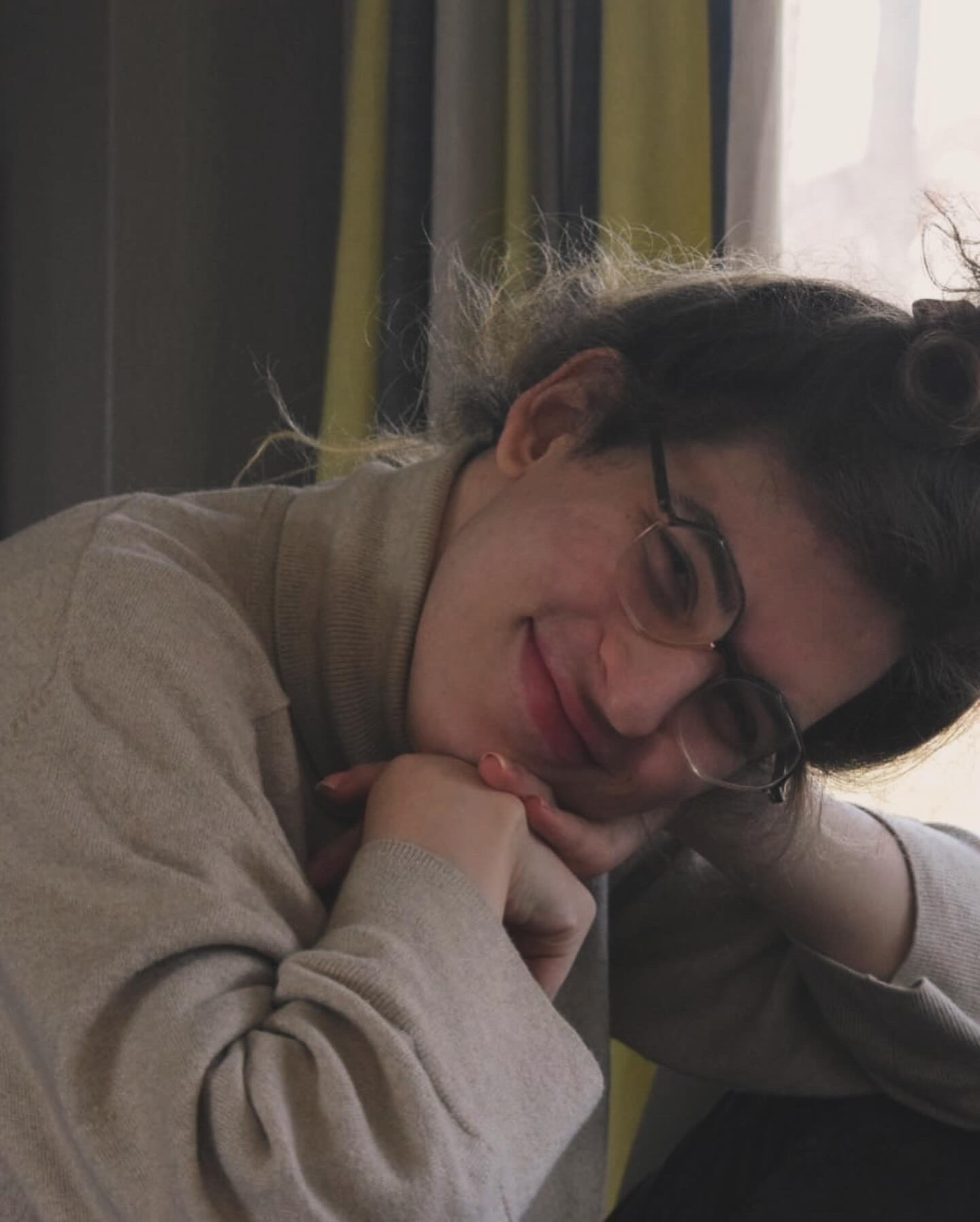 A woman with glasses smiles gently while resting her head on her hands. She is wearing a cozy beige turtleneck sweater, and the soft lighting from a nearby window creates a warm and calm atmosphere. The image conveys a sense of contentment and relaxation.