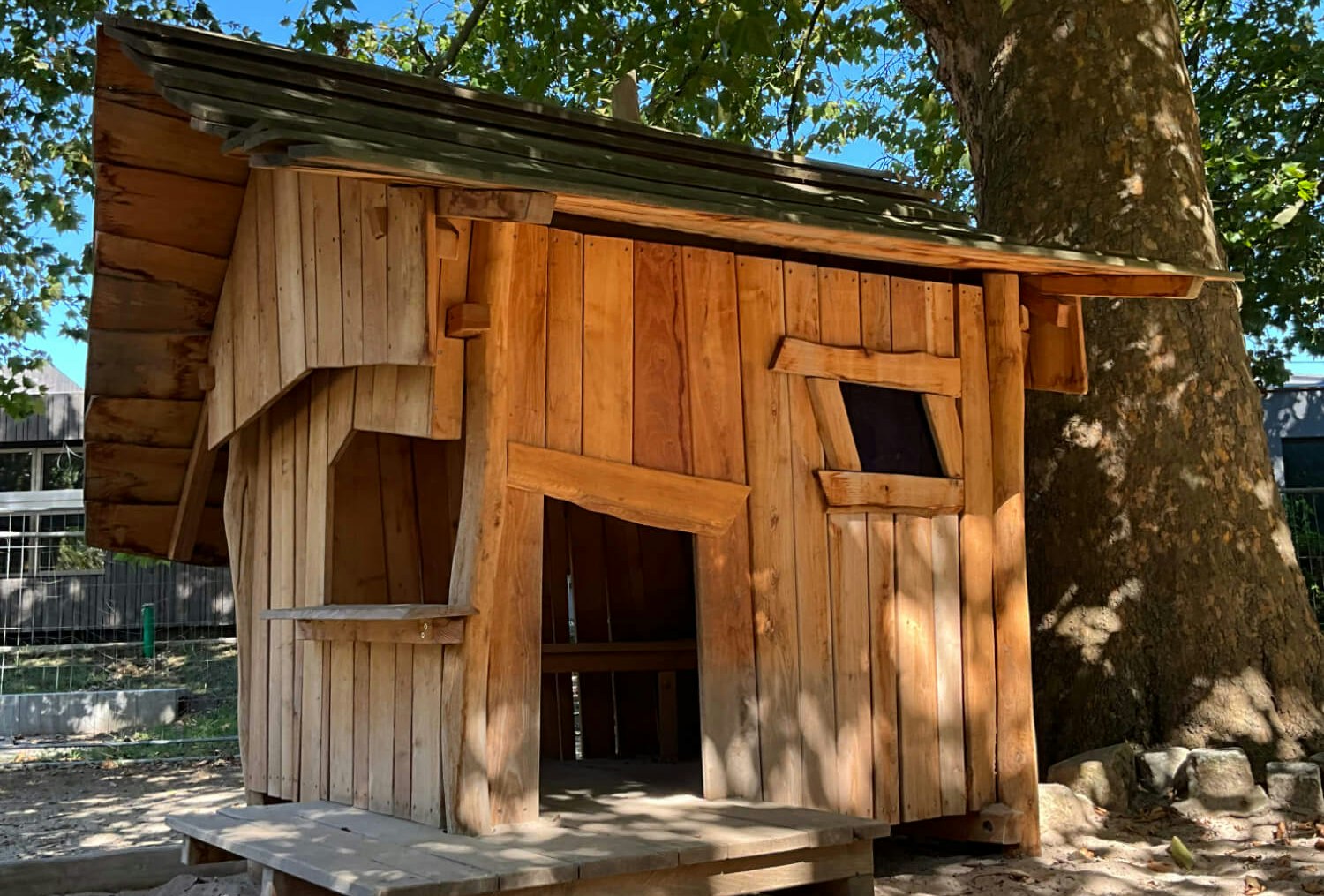 Eine hölzerne Spielhütte im Sand neben einem Baum.
