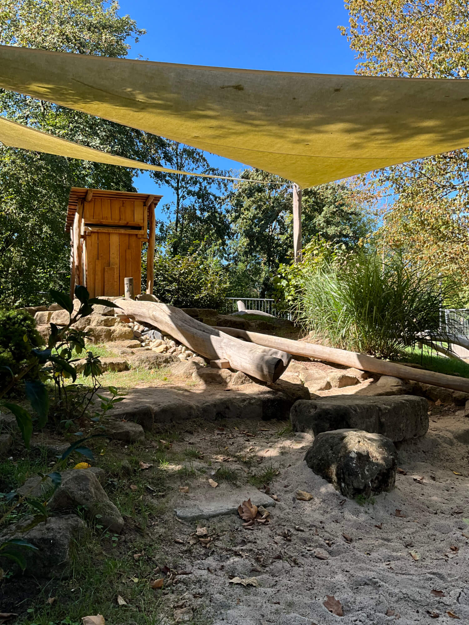 Eine Holzhütte hinter einem Sandkasten mit einem Sonnensegel darüber.