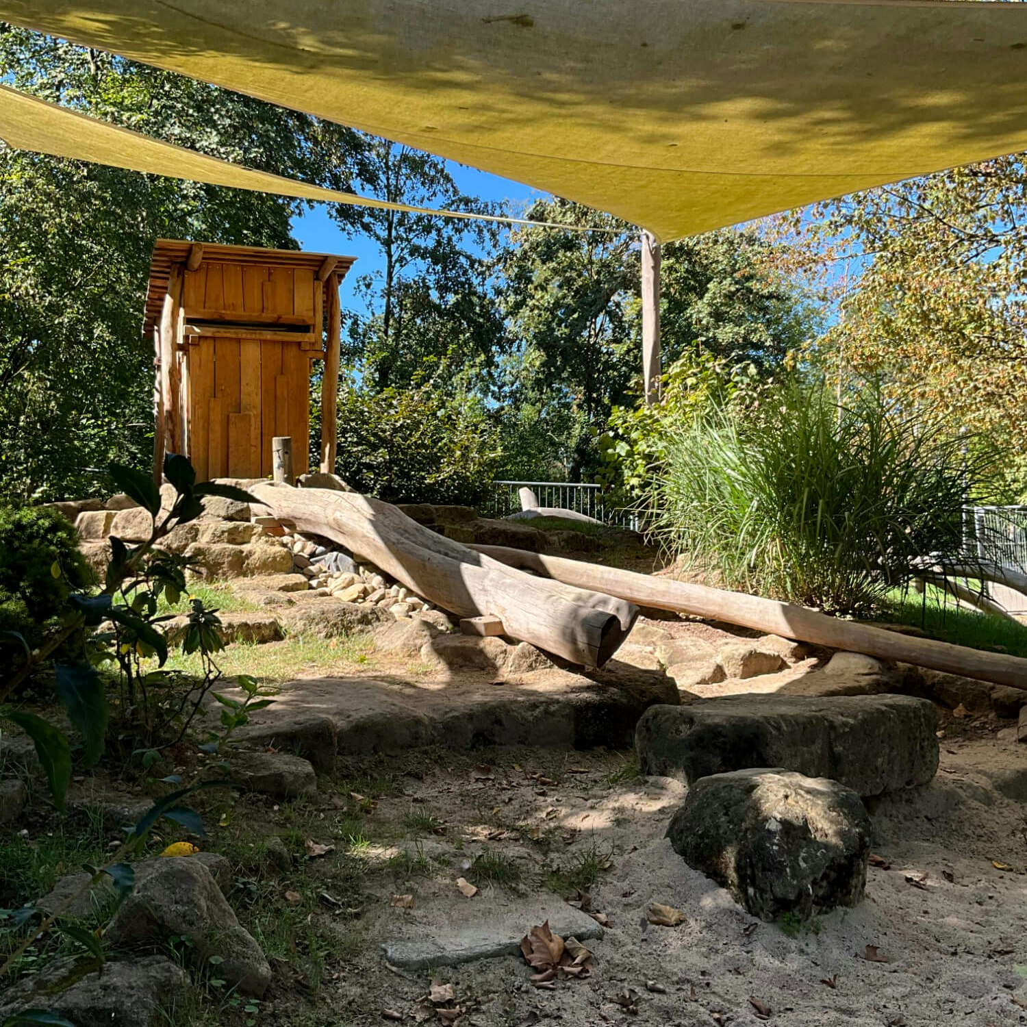 Eine Holzhütte hinter einem Sandkasten mit einem Sonnensegel darüber.