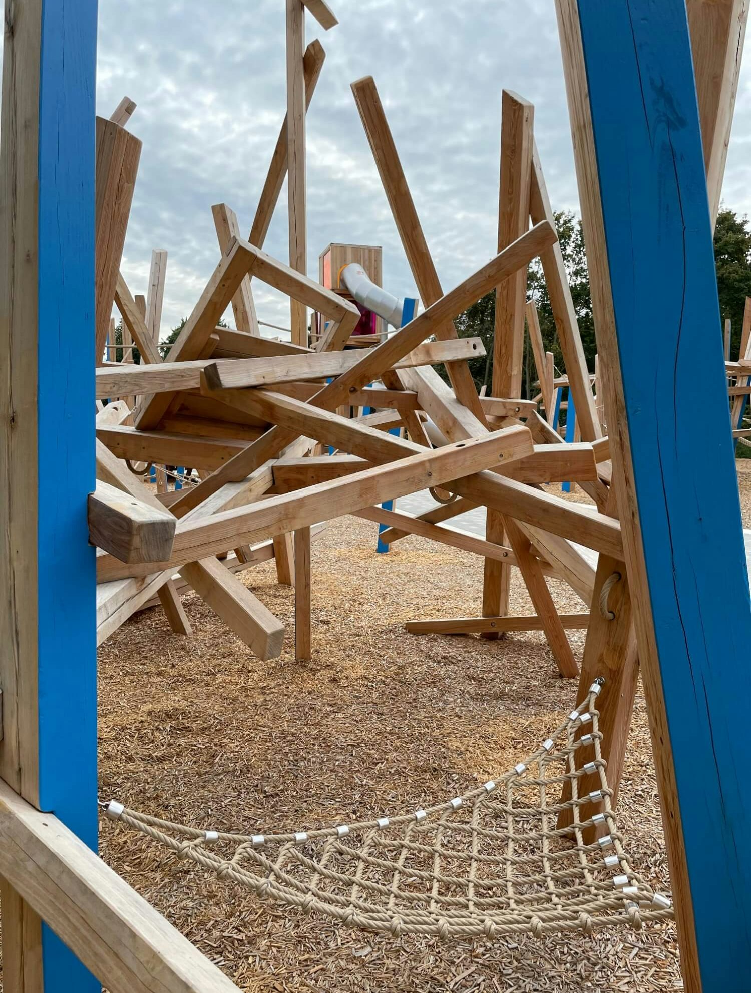 Ein detailreicher Blick auf einen Spielplatz mit komplexen Holzstrukturen. Im Vordergrund sind zwei blaue Holzpfosten zu sehen, zwischen denen sich ein Seilnetz erstreckt. Dahinter befinden sich kunstvoll angeordnete hölzerne Balken, die sich kreuzen und überschneiden. Im Hintergrund ist ein weiteres Spielgerät mit einer Rutsche sichtbar. Der Boden ist mit Holzschnitzeln bedeckt und der Himmel darüber ist bewölkt.