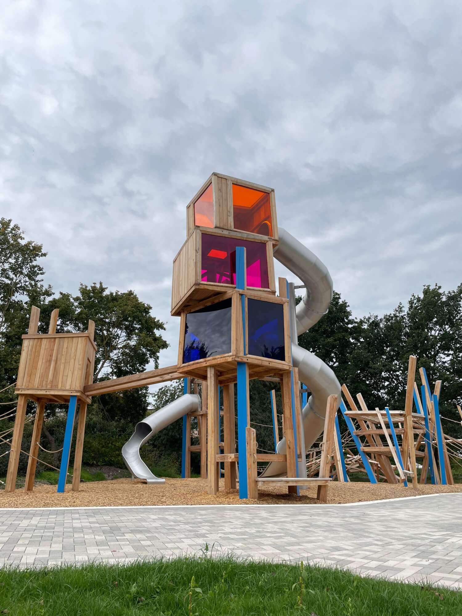 Ein kreativer Holzspielplatz mit mehreren Ebenen und Strukturen. Oben befindet sich eine hölzerne Kabine mit leuchtenden Plexiglasfenstern in Orange, Pink und Blau. Von dieser Kabine führt eine glänzende, silberne Rutsche nach unten. Die Hauptstruktur ist mit blauen Stützen verstärkt und von kleineren Kletterelementen umgeben. Der Untergrund des Spielplatzes ist mit Holzschnitzeln bedeckt, die an einen gepflasterten Gehweg grenzen. Der Himmel darüber ist bewölkt, und am unteren Rand sind Teile einer grünen Wiese sichtbar.
