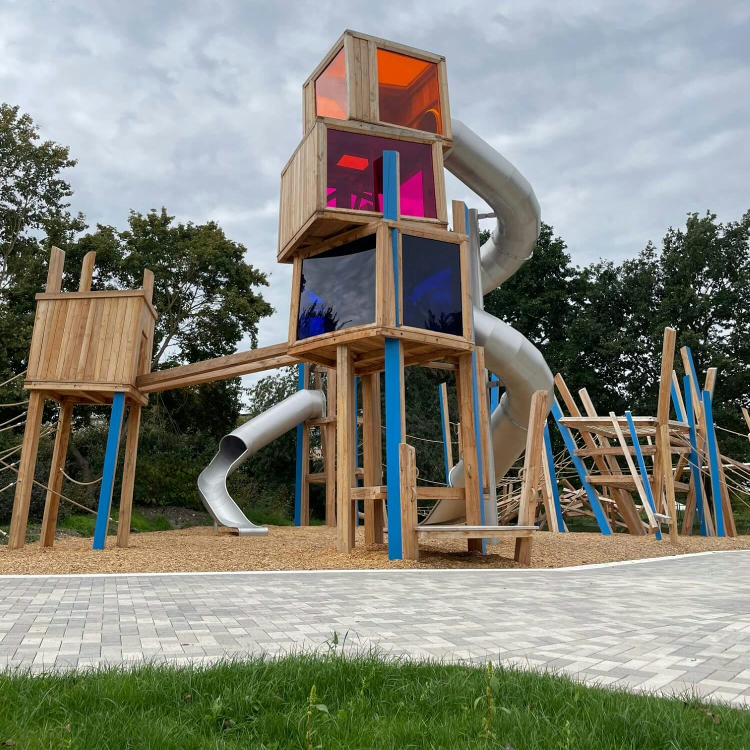 Ein kreativer Holzspielplatz mit mehreren Ebenen und Strukturen. Oben befindet sich eine hölzerne Kabine mit leuchtenden Plexiglasfenstern in Orange, Pink und Blau. Von dieser Kabine führt eine glänzende, silberne Rutsche nach unten. Die Hauptstruktur ist mit blauen Stützen verstärkt und von kleineren Kletterelementen umgeben. Der Untergrund des Spielplatzes ist mit Holzschnitzeln bedeckt, die an einen gepflasterten Gehweg grenzen. Der Himmel darüber ist bewölkt, und am unteren Rand sind Teile einer grünen Wiese sichtbar.