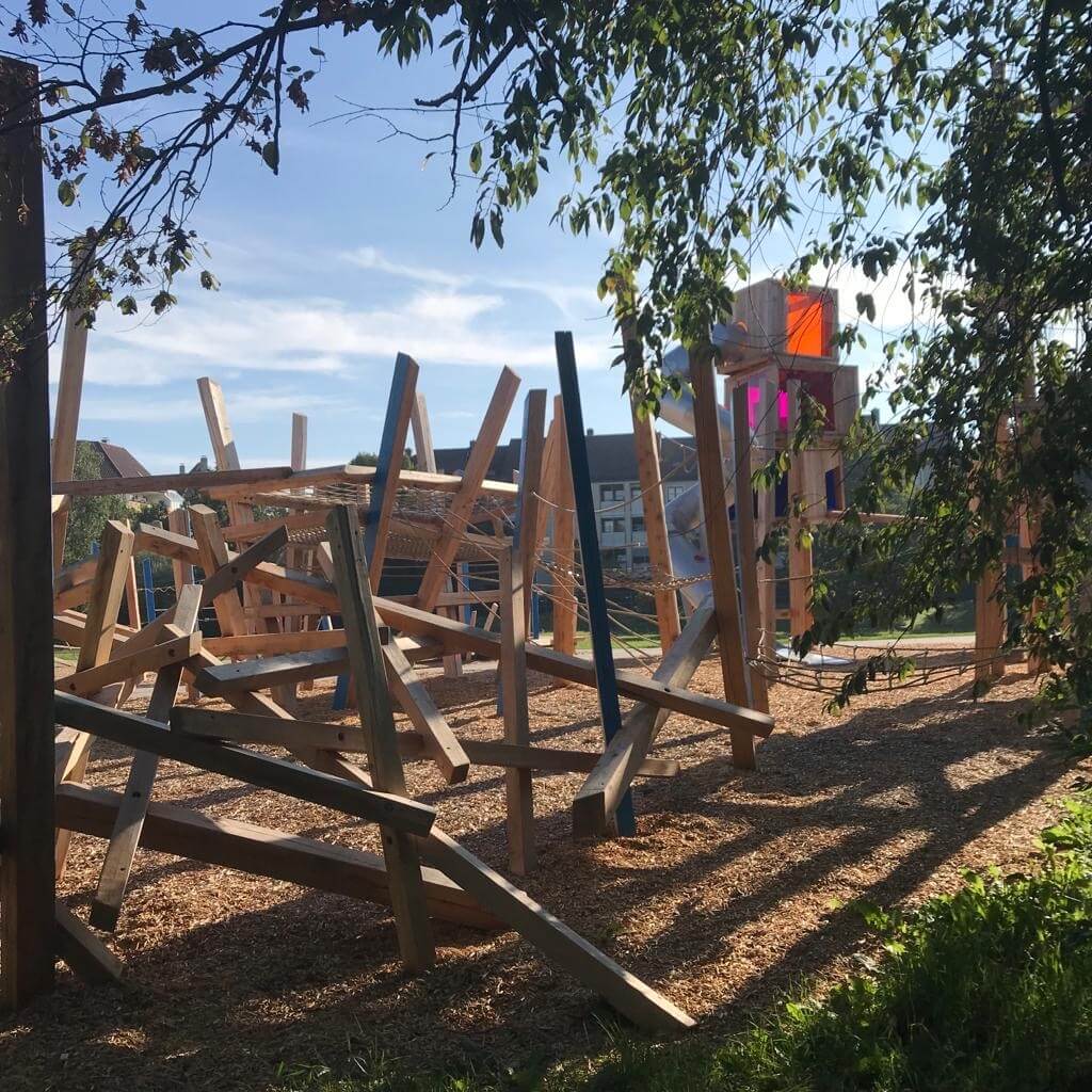 Ein moderner Spielplatz mit vielseitigen hölzernen Kletterstrukturen, die durch Balken und Seile verbunden sind. Auf der rechten Seite steht ein größeres Holzspielgerät mit bunten Plexiglasfenstern in Orange und Pink. Der gesamte Bereich ist mit Holzschnitzeln bedeckt. Grüne Bäume und Sträucher rahmen den Vordergrund ein, während im Hintergrund Stadthäuser unter einem blauen Himmel zu sehen sind. Das Sonnenlicht betont die Strukturen und lässt Schatten auf den Boden fallen.