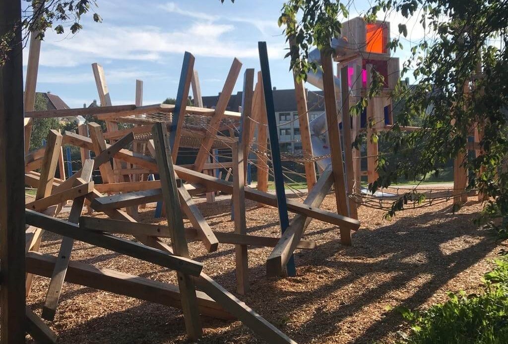 Ein moderner Spielplatz mit vielseitigen hölzernen Kletterstrukturen, die durch Balken und Seile verbunden sind. Auf der rechten Seite steht ein größeres Holzspielgerät mit bunten Plexiglasfenstern in Orange und Pink. Der gesamte Bereich ist mit Holzschnitzeln bedeckt. Grüne Bäume und Sträucher rahmen den Vordergrund ein, während im Hintergrund Stadthäuser unter einem blauen Himmel zu sehen sind. Das Sonnenlicht betont die Strukturen und lässt Schatten auf den Boden fallen.