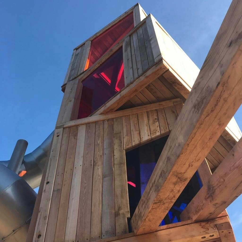 Detailansicht einer hölzernen Spielplatzstruktur. Oben ist eine Kabine mit farbigen Plexiglasfenstern in Rot und Pink zu sehen, darunter befindet sich ein weiteres Fenster in Blau. Die Holzplanken der Kabine sind rustikal und verwittert, und es gibt sichtbare Holzträger, die die Struktur stützen. Ein Abschnitt einer silbernen Rutsche ist am linken Rand sichtbar. Der Hintergrund zeigt einen klaren blauen Himmel.