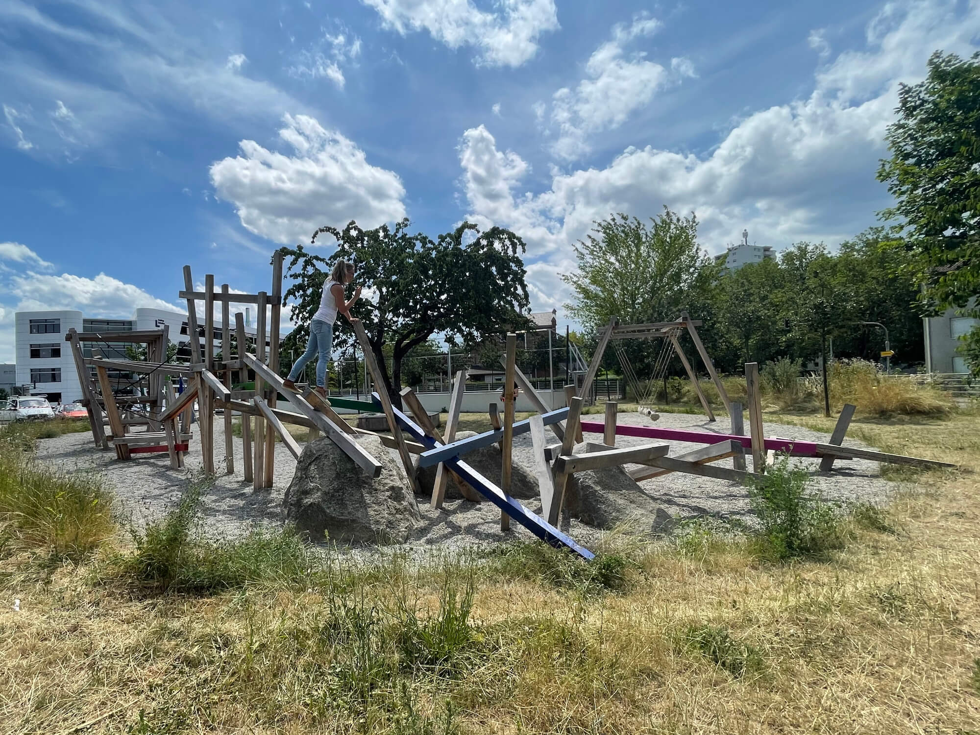 Ein sonniger Spielplatz mit trockenem Gras im Vordergrund und mehreren Holzspielgeräten in verschiedenen Farben. Eine Person steht auf einem der Spielgeräte und balanciert. Im Hintergrund sind moderne Gebäude und ein blauer Himmel mit Wolken zu sehen.