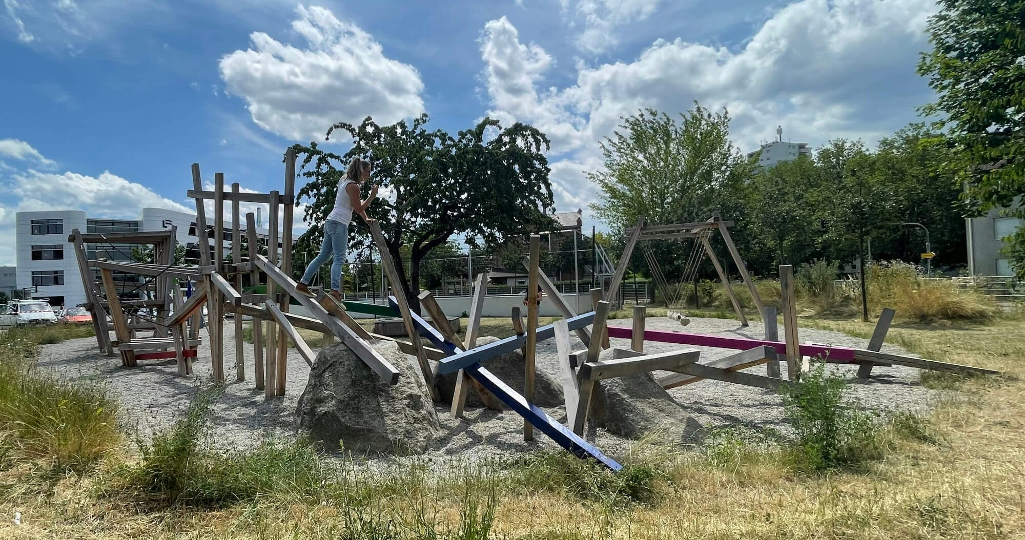 Ein sonniger Spielplatz mit trockenem Gras im Vordergrund und mehreren Holzspielgeräten in verschiedenen Farben. Eine Person steht auf einem der Spielgeräte und balanciert. Im Hintergrund sind moderne Gebäude und ein blauer Himmel mit Wolken zu sehen.