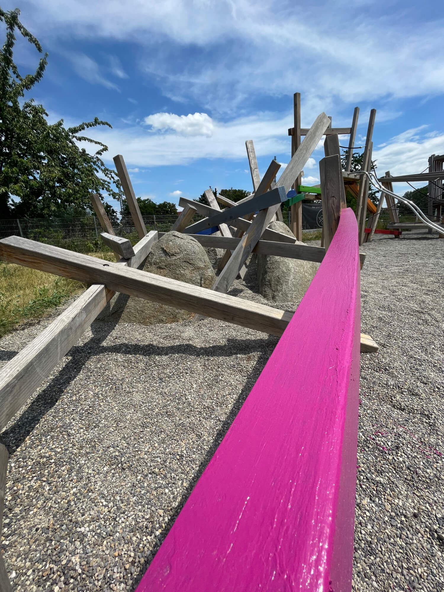 Detailaufnahme eines Kies-Spielplatzes mit einem frisch in Pink gestrichenen Holzbalken im Vordergrund. Dahinter sind diverse Holzkonstruktionen, Steine und Spielgeräte zu erkennen, alles unter einem klaren blauen Himmel.