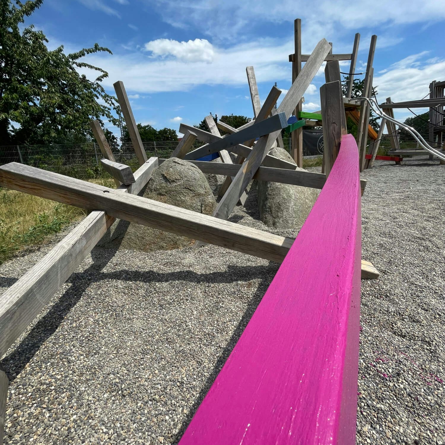 Detailaufnahme eines Kies-Spielplatzes mit einem frisch in Pink gestrichenen Holzbalken im Vordergrund. Dahinter sind diverse Holzkonstruktionen, Steine und Spielgeräte zu erkennen, alles unter einem klaren blauen Himmel.
