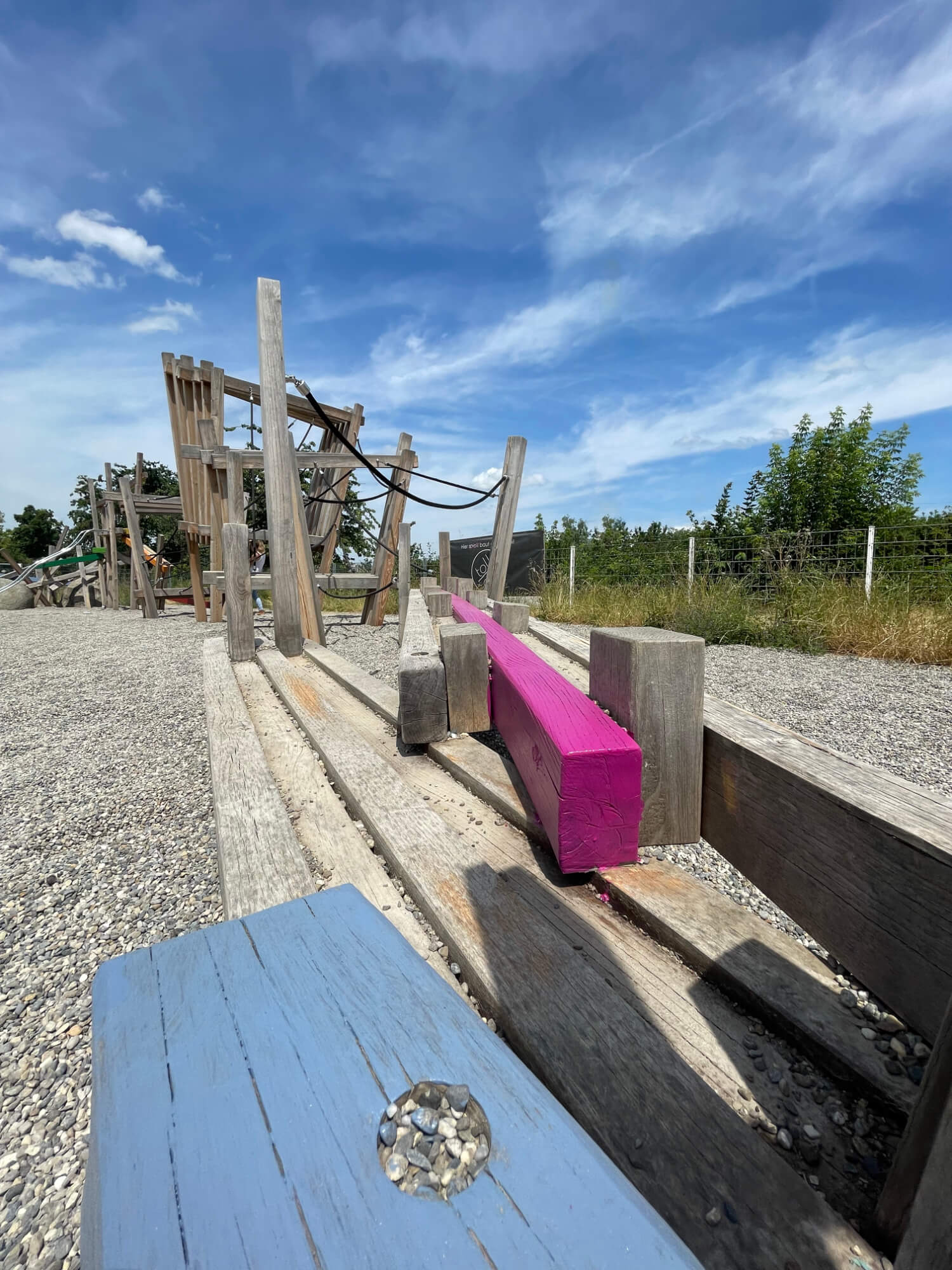 Detailansicht eines Kies-Spielplatzes mit mehreren Holzbalken. Einige Balken sind frisch in leuchtenden Farben wie Blau und Pink gestrichen, während andere noch unbehandelt sind. Im Hintergrund sind Spielgeräte und Bäume unter einem blauen Himmel mit Wolken zu sehen.