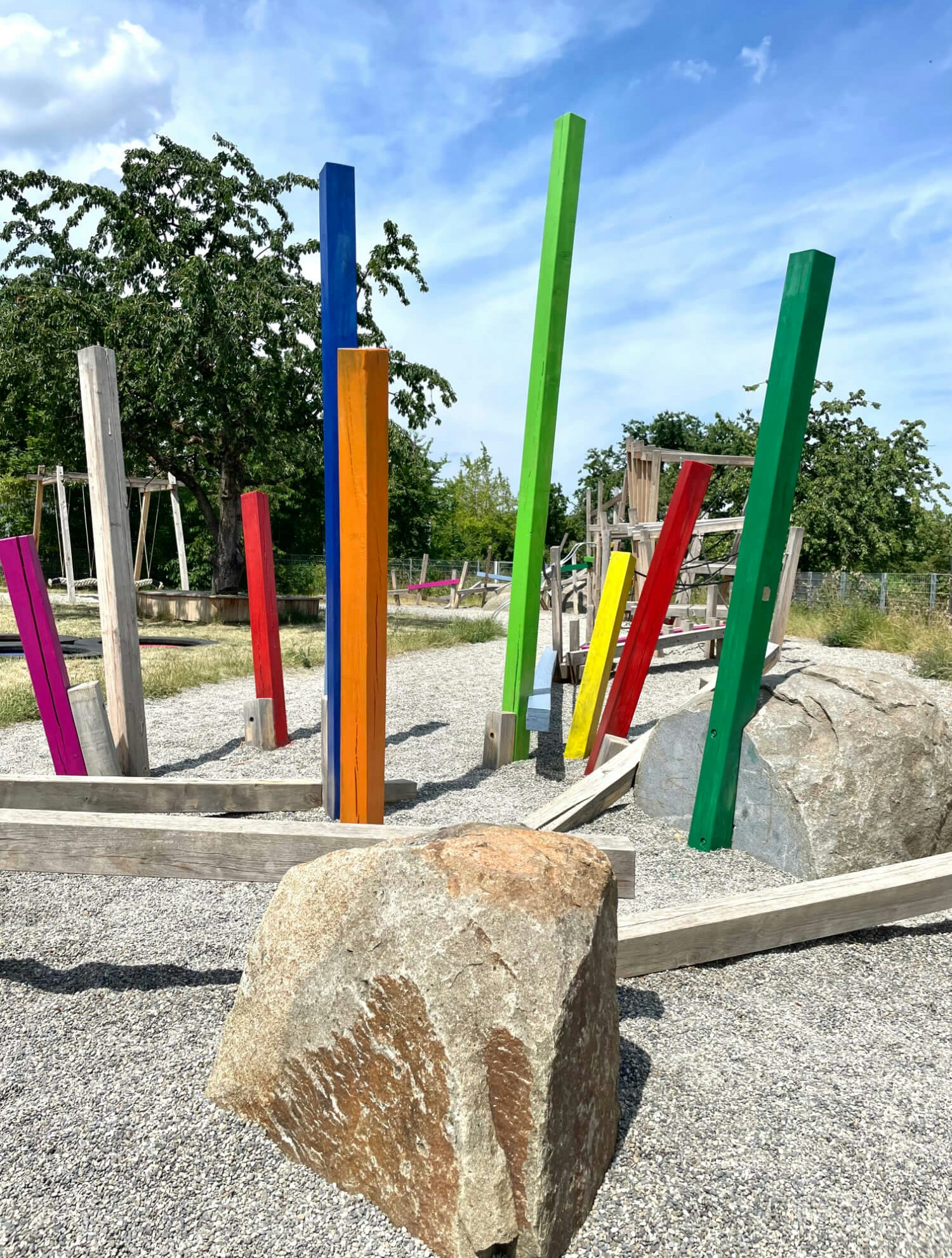 Farbige vertikale Holzbalken unterschiedlicher Höhe auf einem Spielplatz mit Kiesboden, Bäumen im Hintergrund und großen Steinen im Vordergrund.