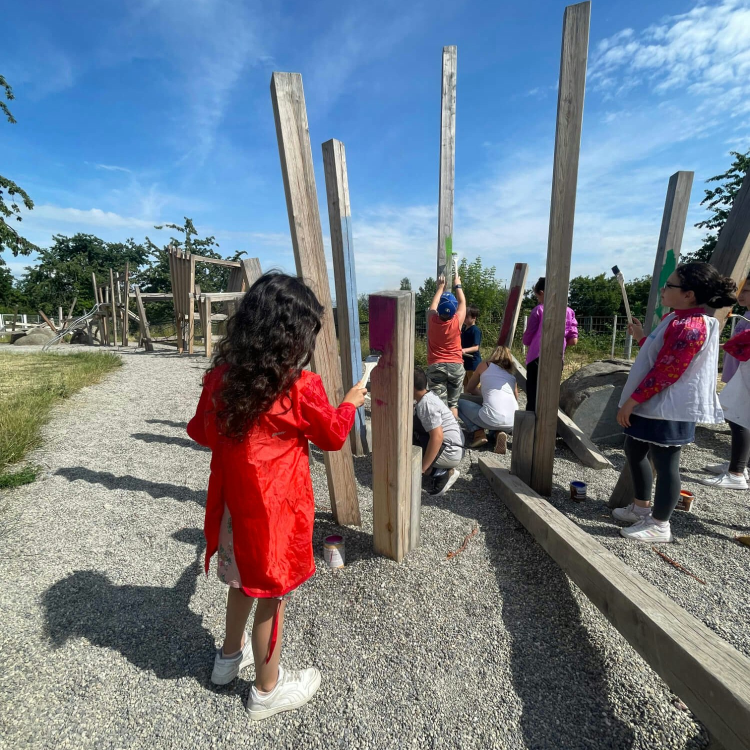 Kinder und Erwachsene streichen vertikale Holzbalken auf einem Kies-Spielplatz. Ein Mädchen im Vordergrund mit einem roten Mantel hält einen Pinsel. Im Hintergrund sind Bäume und ein blauer Himmel zu sehen.