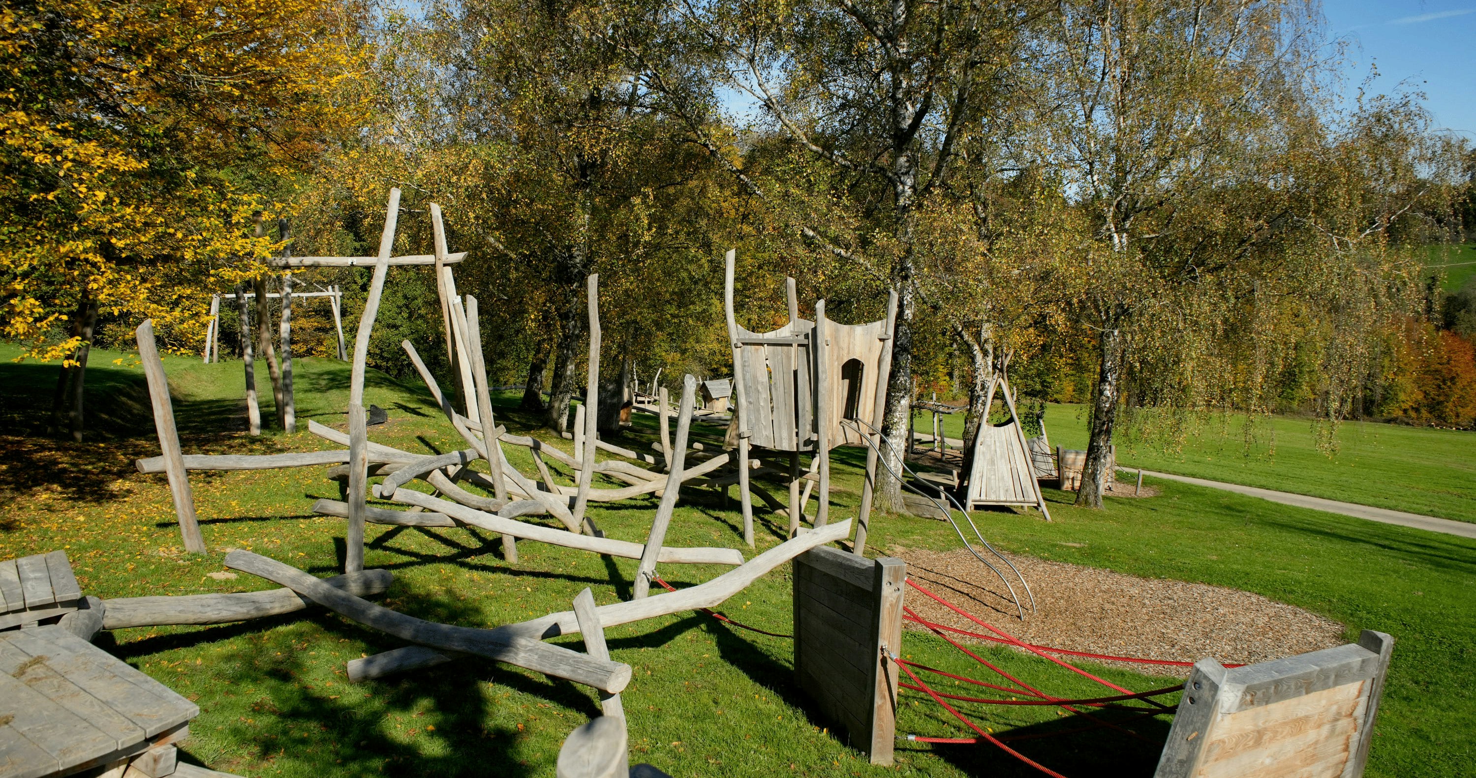 Zwei dreieckige Holzkonstruktionen, die an Tipis erinnern, stehen auf einer Rasenfläche, umgeben von Bäumen mit Herbstlaub.
