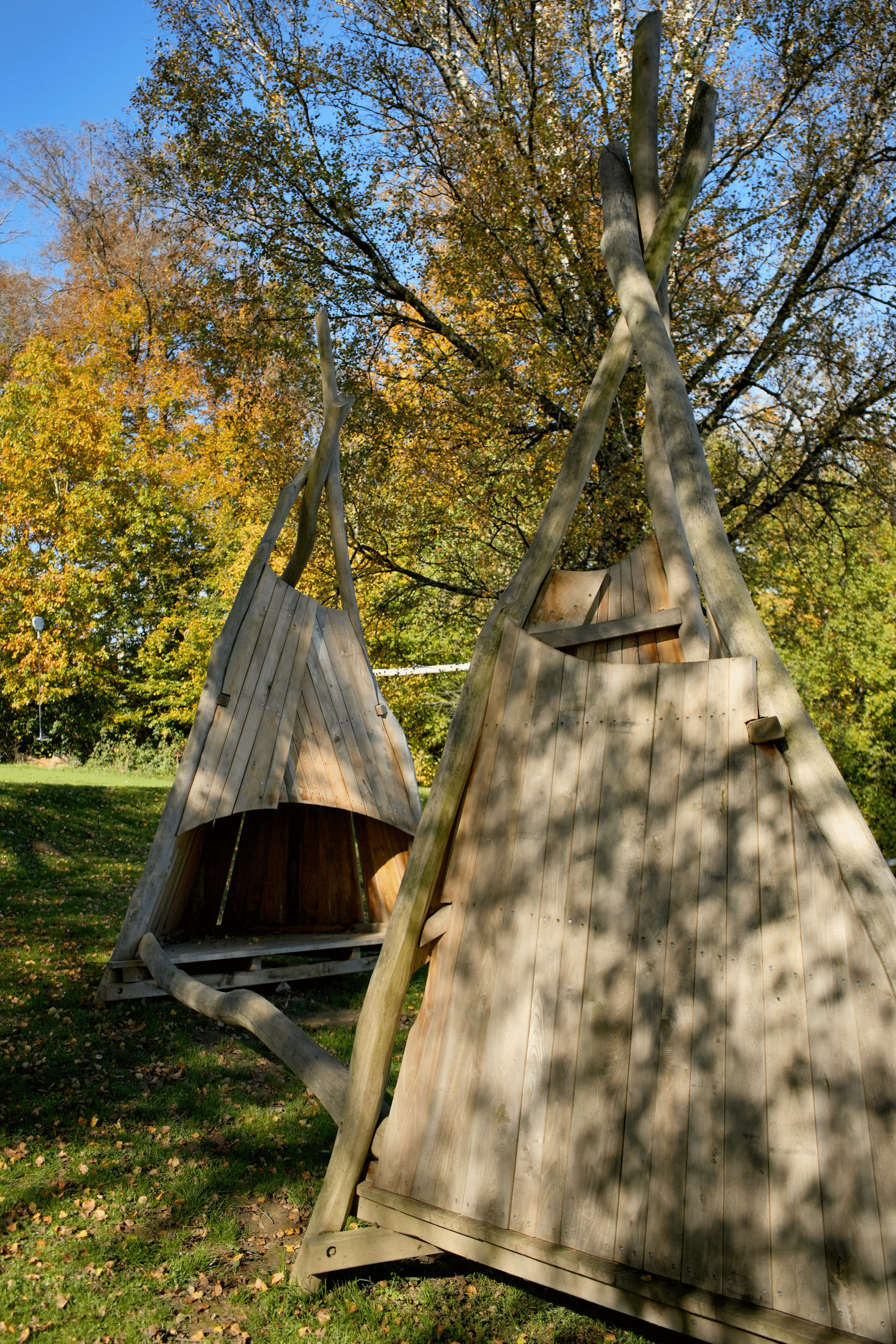 An einem sonnigen Tag stehen zwei dreieckige Holzkonstruktionen, die an Tipis erinnern, auf Gras, im Hintergrund sind Bäume zu sehen.