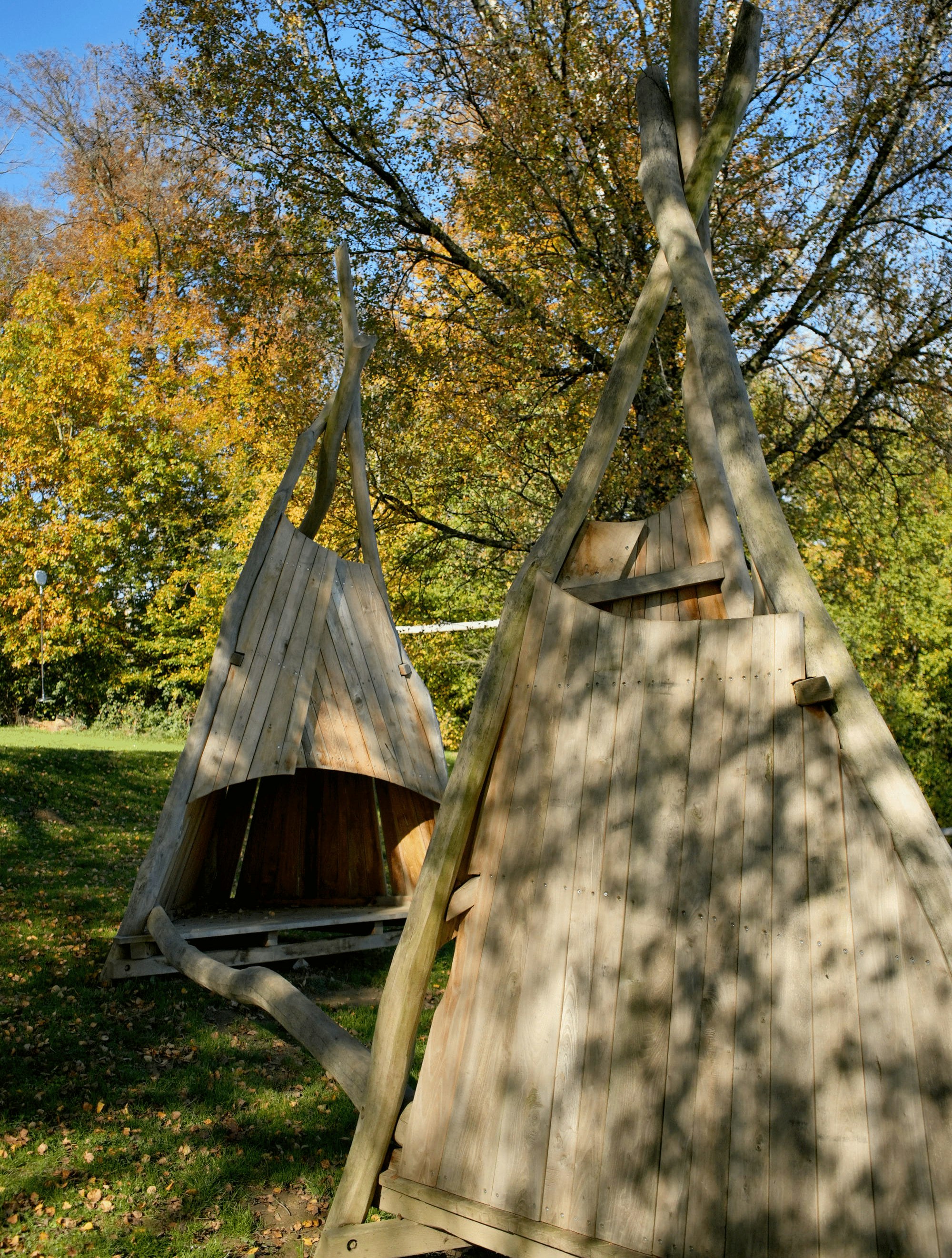 An einem sonnigen Tag stehen zwei dreieckige Holzkonstruktionen, die an Tipis erinnern, auf Gras, im Hintergrund sind Bäume zu sehen.