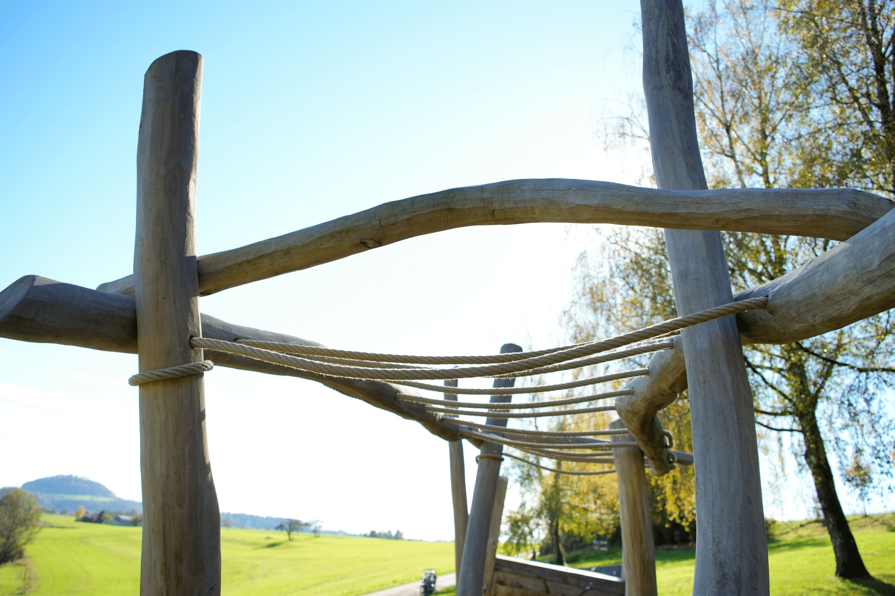 Hölzernes Klettergerüst mit Seilen auf einem Spielplatz vor dem Hintergrund eines klaren blauen Himmels und einer grasbewachsenen Landschaft.