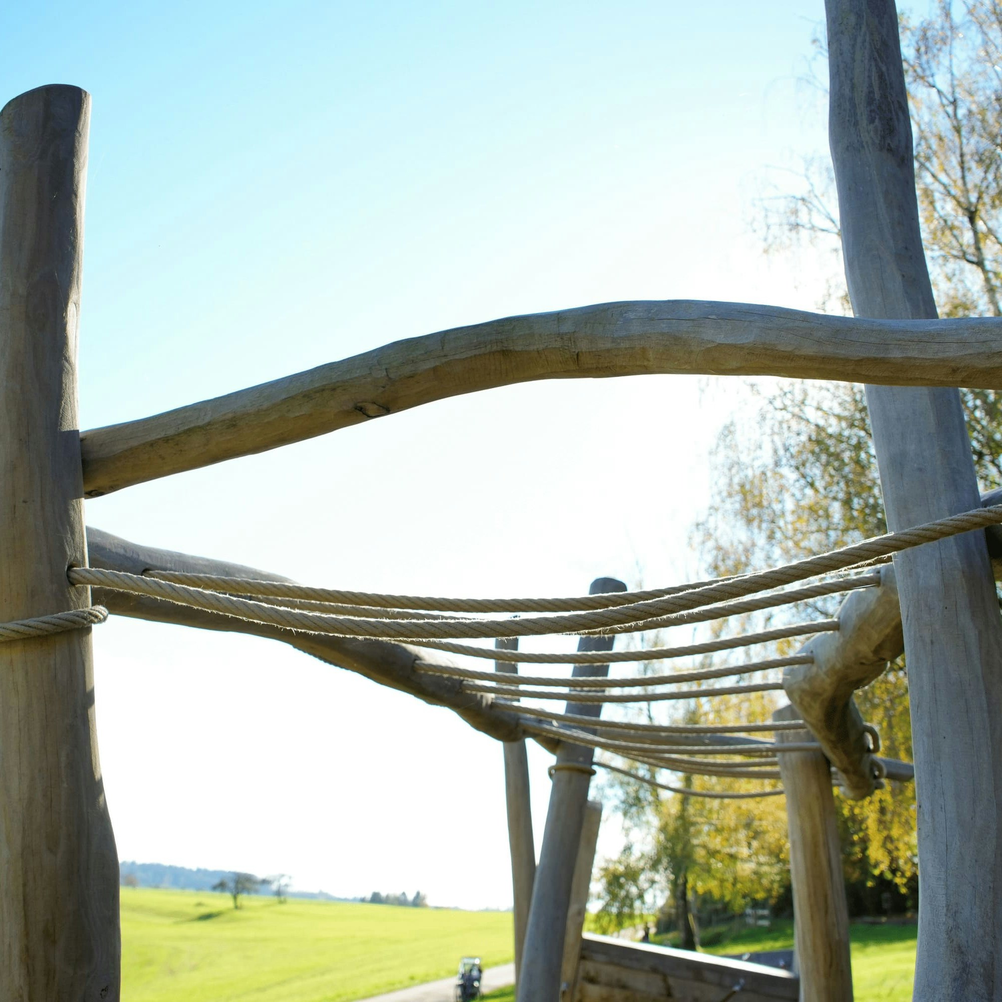 Hölzernes Klettergerüst mit Seilen auf einem Spielplatz vor dem Hintergrund eines klaren blauen Himmels und einer grasbewachsenen Landschaft.