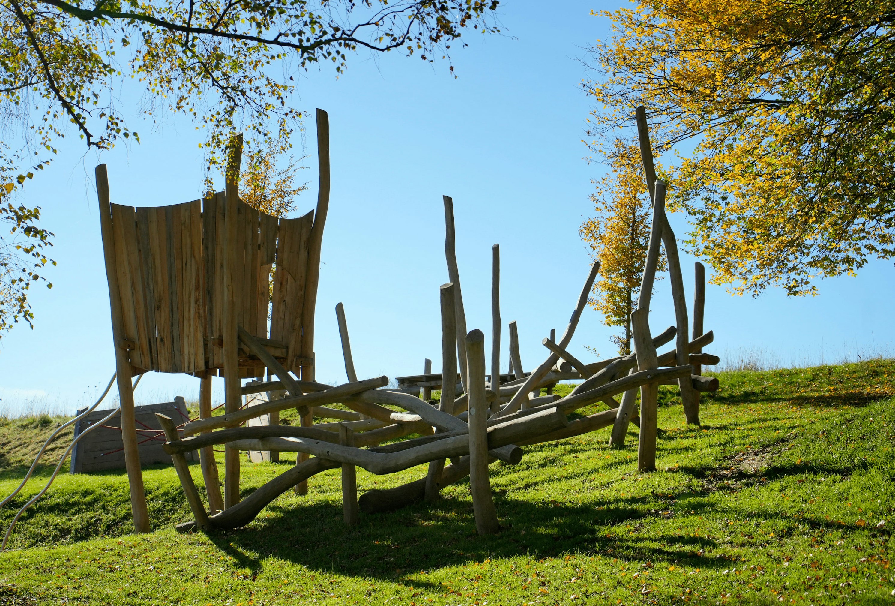 Hölzerne Spielplatzstruktur mit Rutsche und Kletterelementen auf einer Rasenfläche. Umliegende Bäume haben Herbstlaub unter einem klaren blauen Himmel.