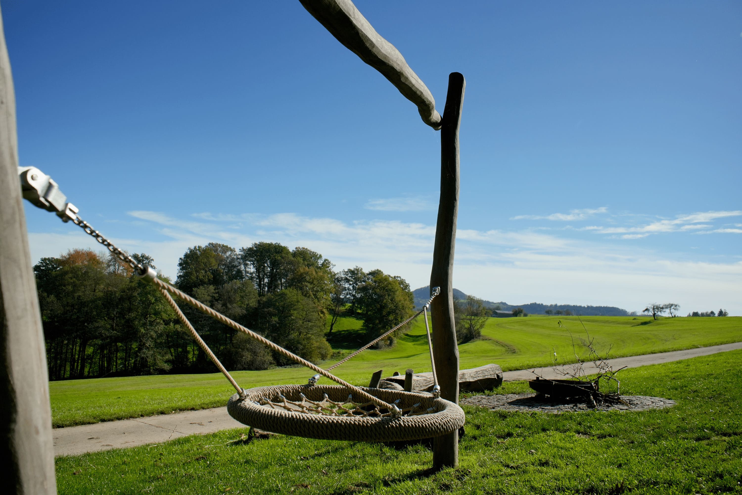 Eine runde Schaukel hängt an Holzpfosten auf einer Wiese, mit einem gewundenen Pfad und Bäumen im Hintergrund unter einem klaren blauen Himmel.