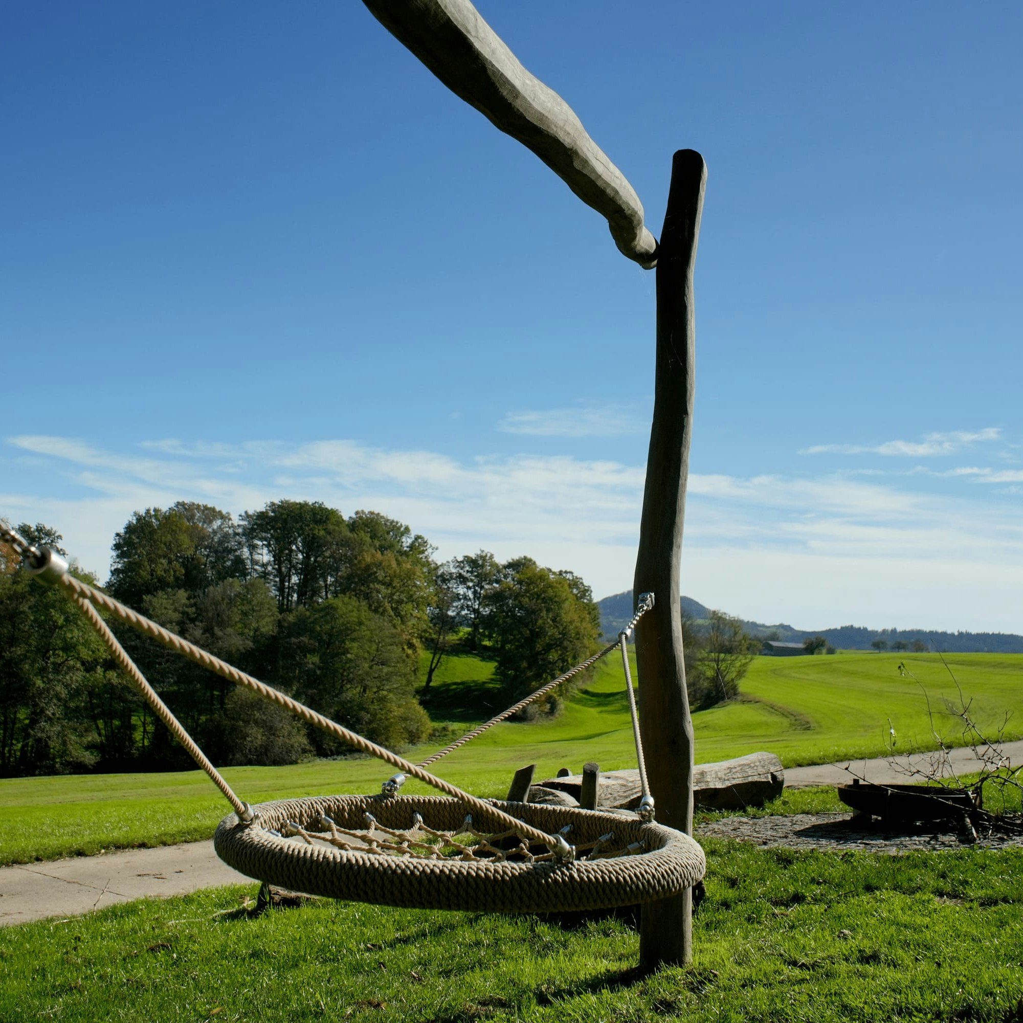 Eine runde Schaukel hängt an Holzpfosten auf einer Wiese, mit einem gewundenen Pfad und Bäumen im Hintergrund unter einem klaren blauen Himmel.