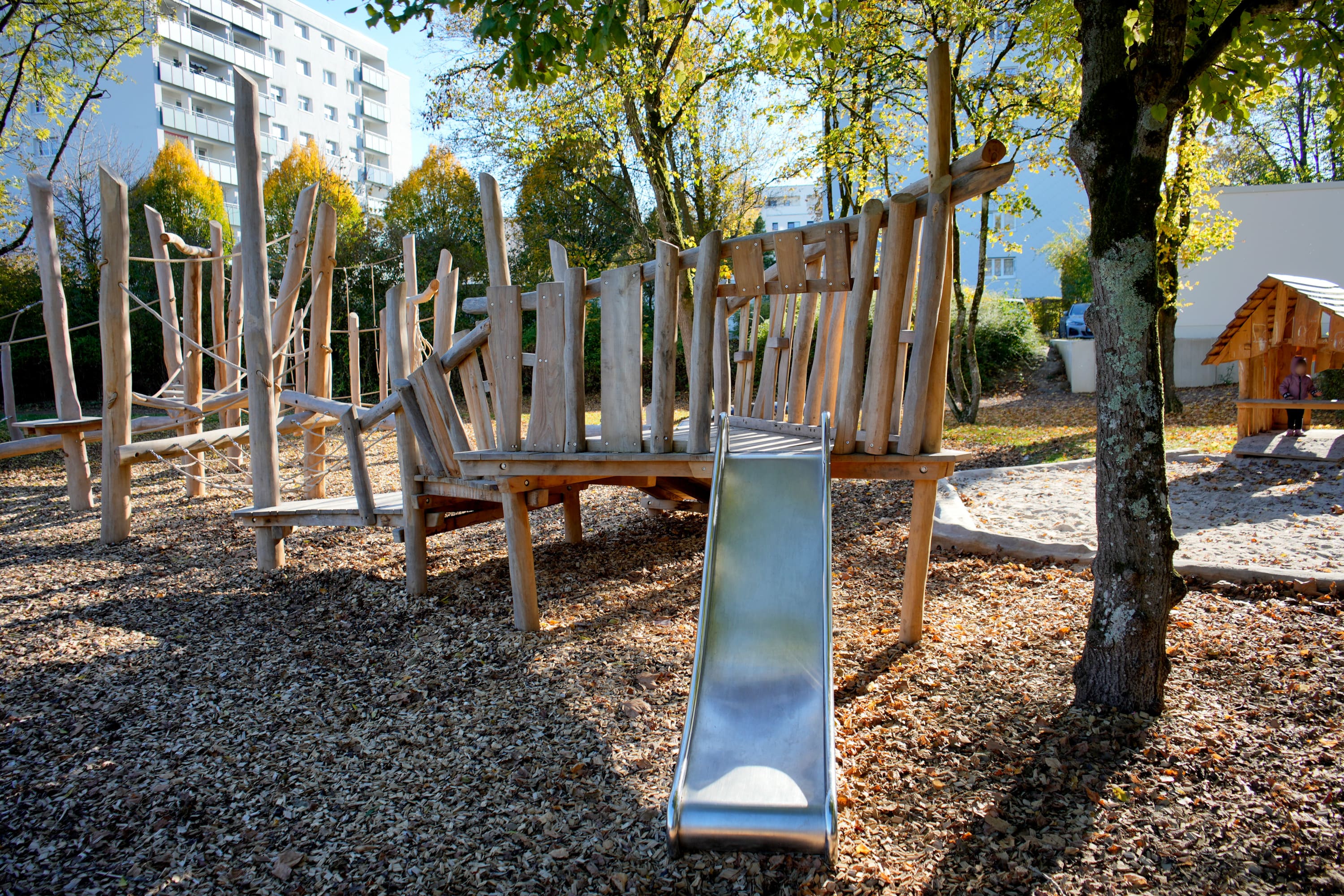 Ein hölzernes Spielplatzgerüst mit einer Metallrutsche ist von Bäumen und Rindenmulch umgeben. Im Hintergrund ist ein Wohnhaus zu sehen.