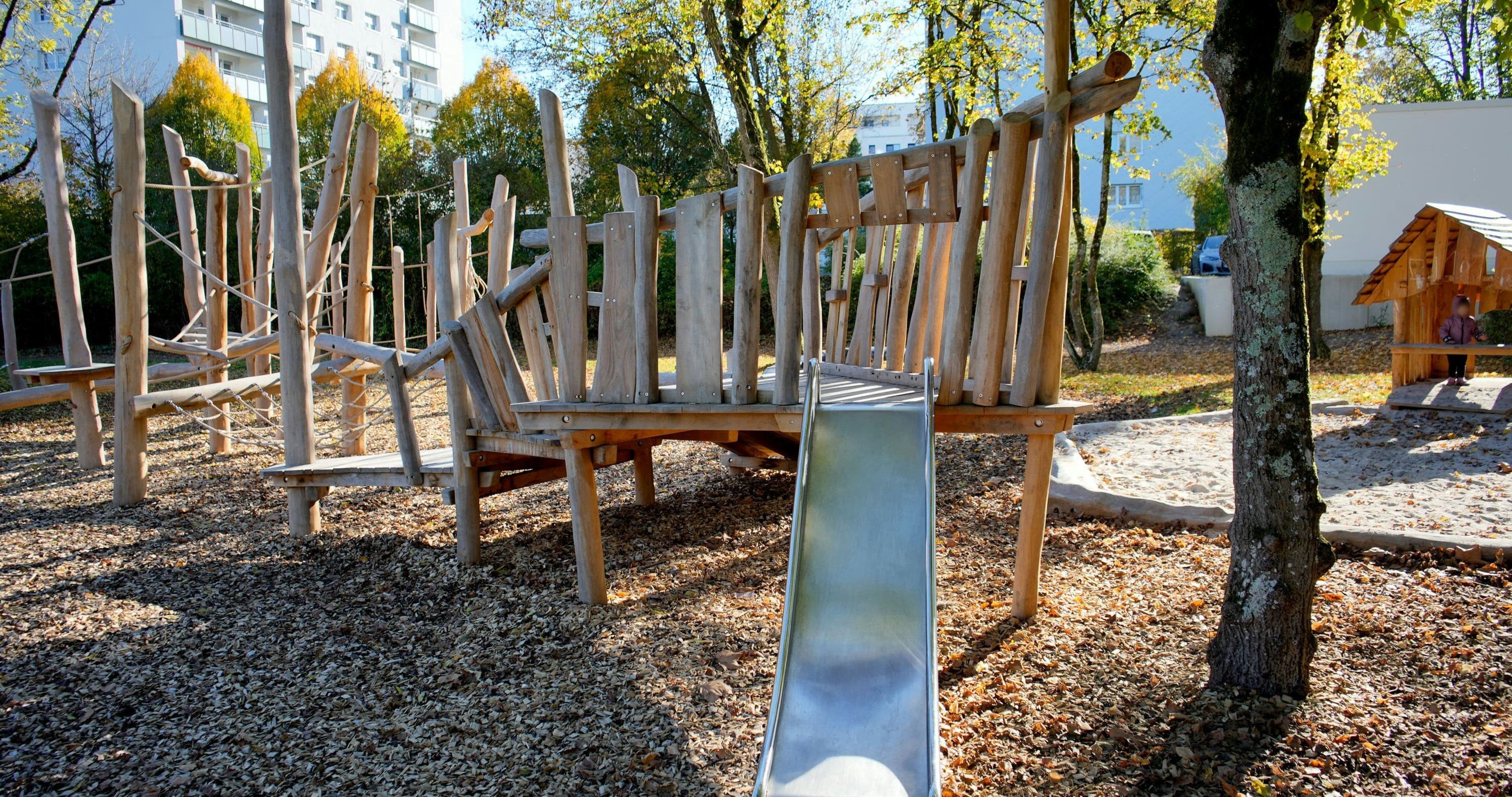 Ein hölzernes Spielplatzgerüst mit einer Metallrutsche ist von Bäumen und Rindenmulch umgeben. Im Hintergrund ist ein Wohnhaus zu sehen.