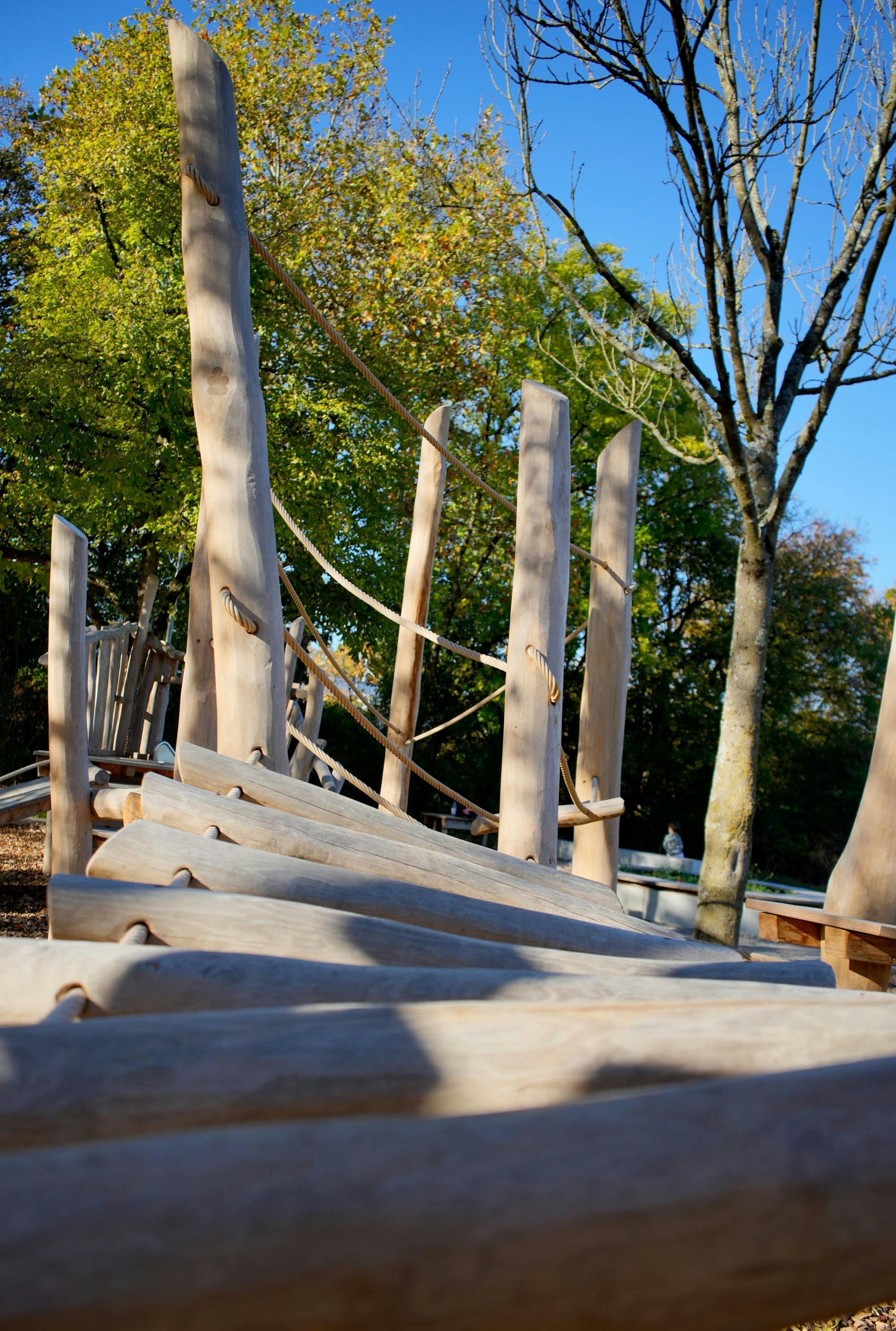 Hölzernes Klettergerüst mit vertikalen Pfosten und Seilen auf einem Spielplatz, umgeben von Bäumen und klarem blauen Himmel.