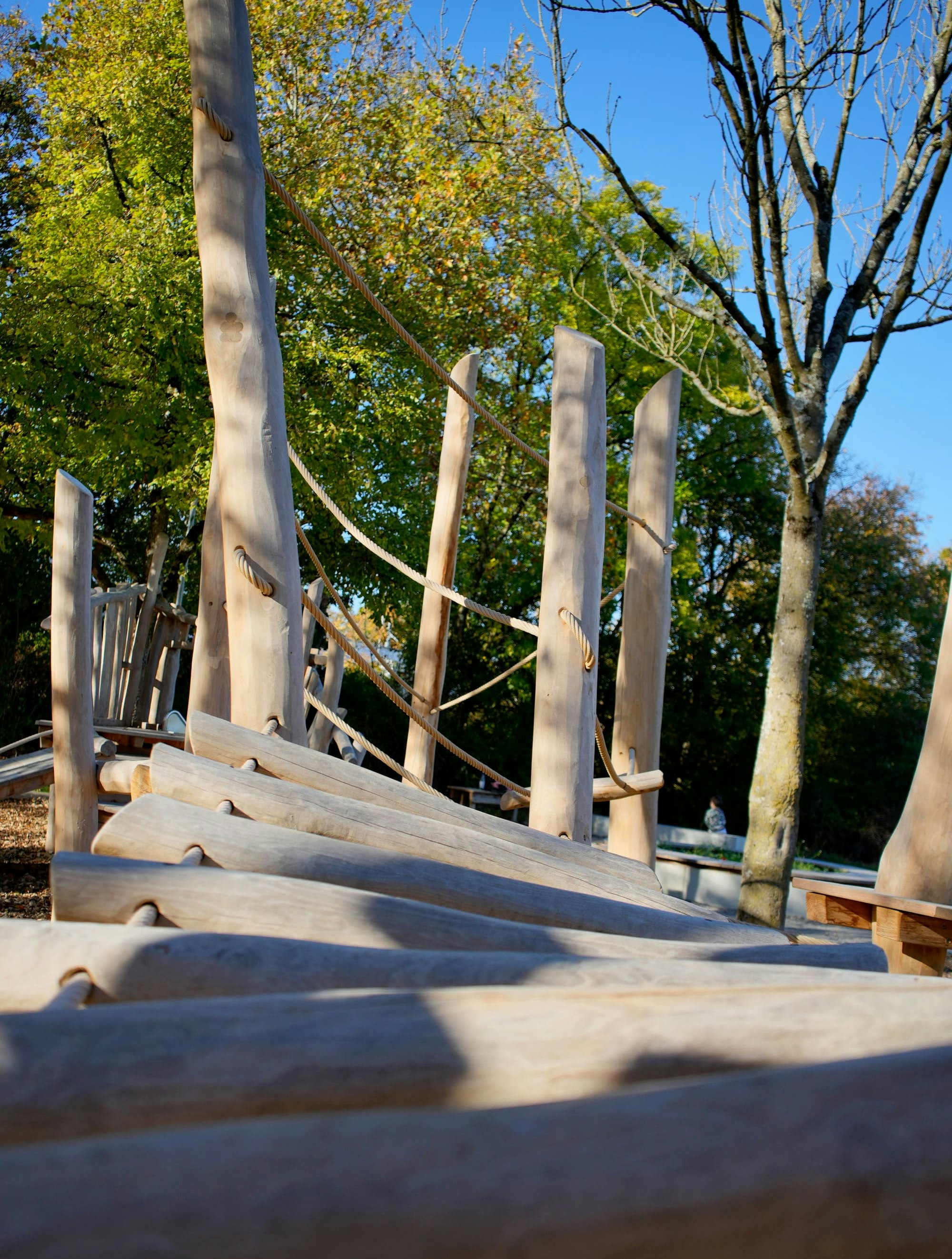 Hölzernes Klettergerüst mit vertikalen Pfosten und Seilen auf einem Spielplatz, umgeben von Bäumen und klarem blauen Himmel.