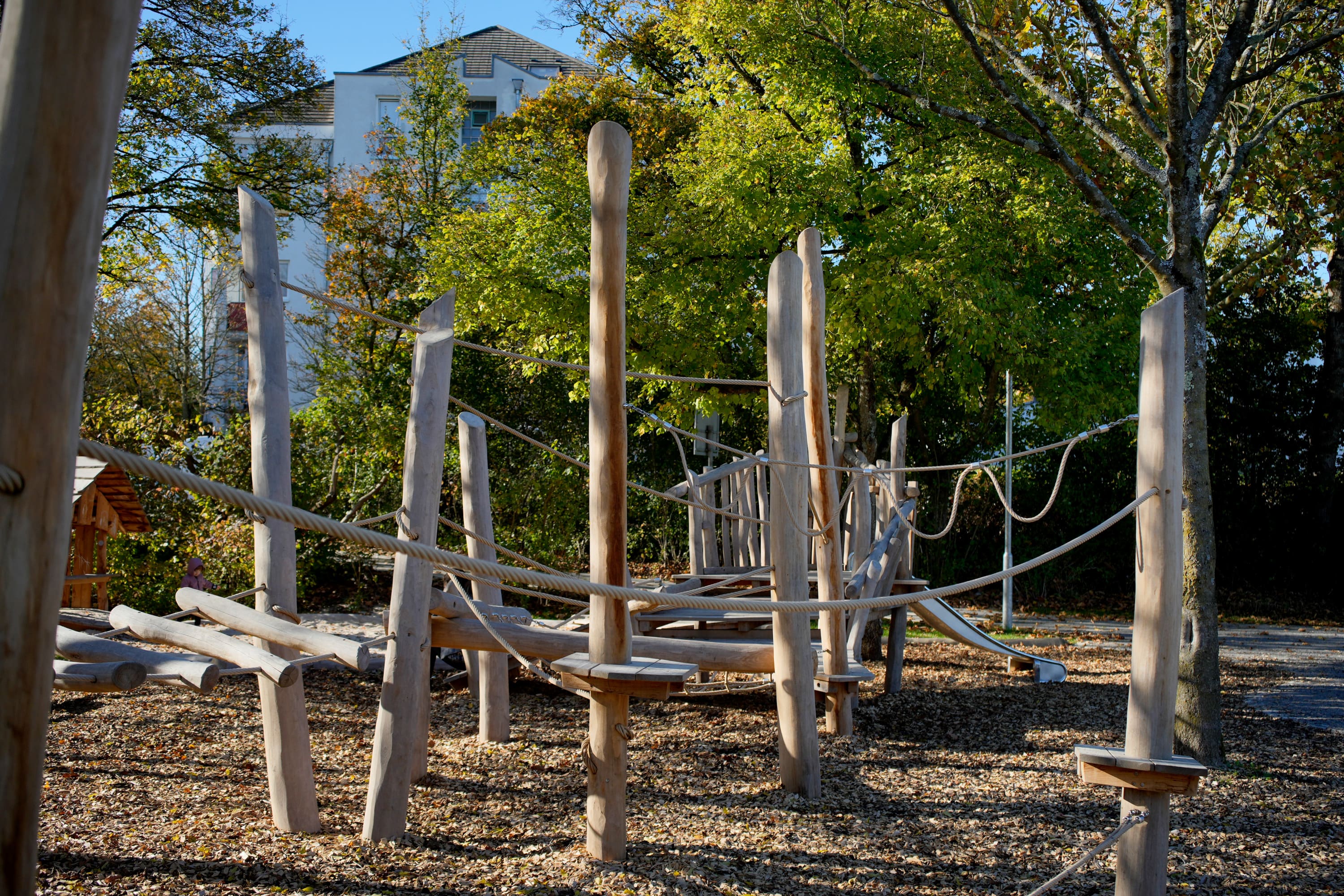 Holzspielplatz mit Kletterseilen und -stangen, umgeben von Bäumen, unter klarem Himmel.