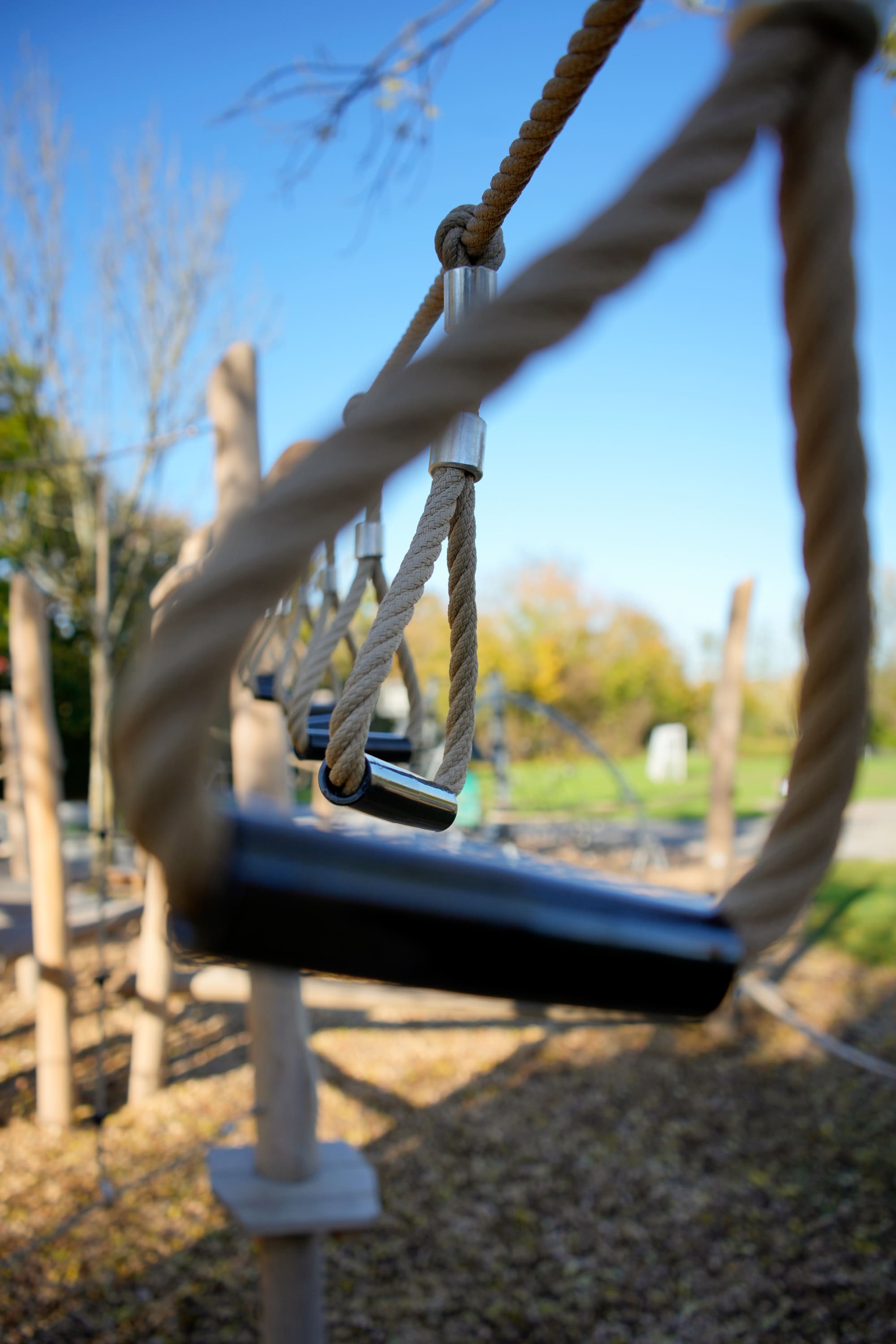 Nahaufnahme eines Seilhindernisparcours auf einem Spielplatz im Freien mit dicken Seilen und Holzgriffen. Im Hintergrund sind ein klarer blauer Himmel und verschwommene Bäume zu sehen.