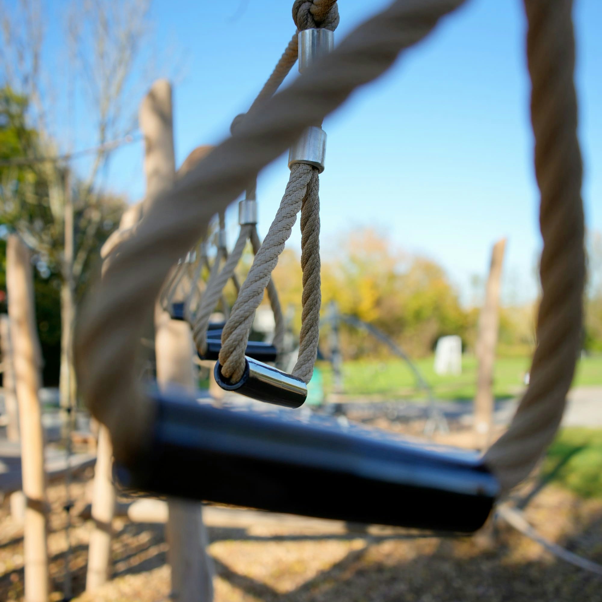 Nahaufnahme eines Seilhindernisparcours auf einem Spielplatz im Freien mit dicken Seilen und Holzgriffen. Im Hintergrund sind ein klarer blauer Himmel und verschwommene Bäume zu sehen.
