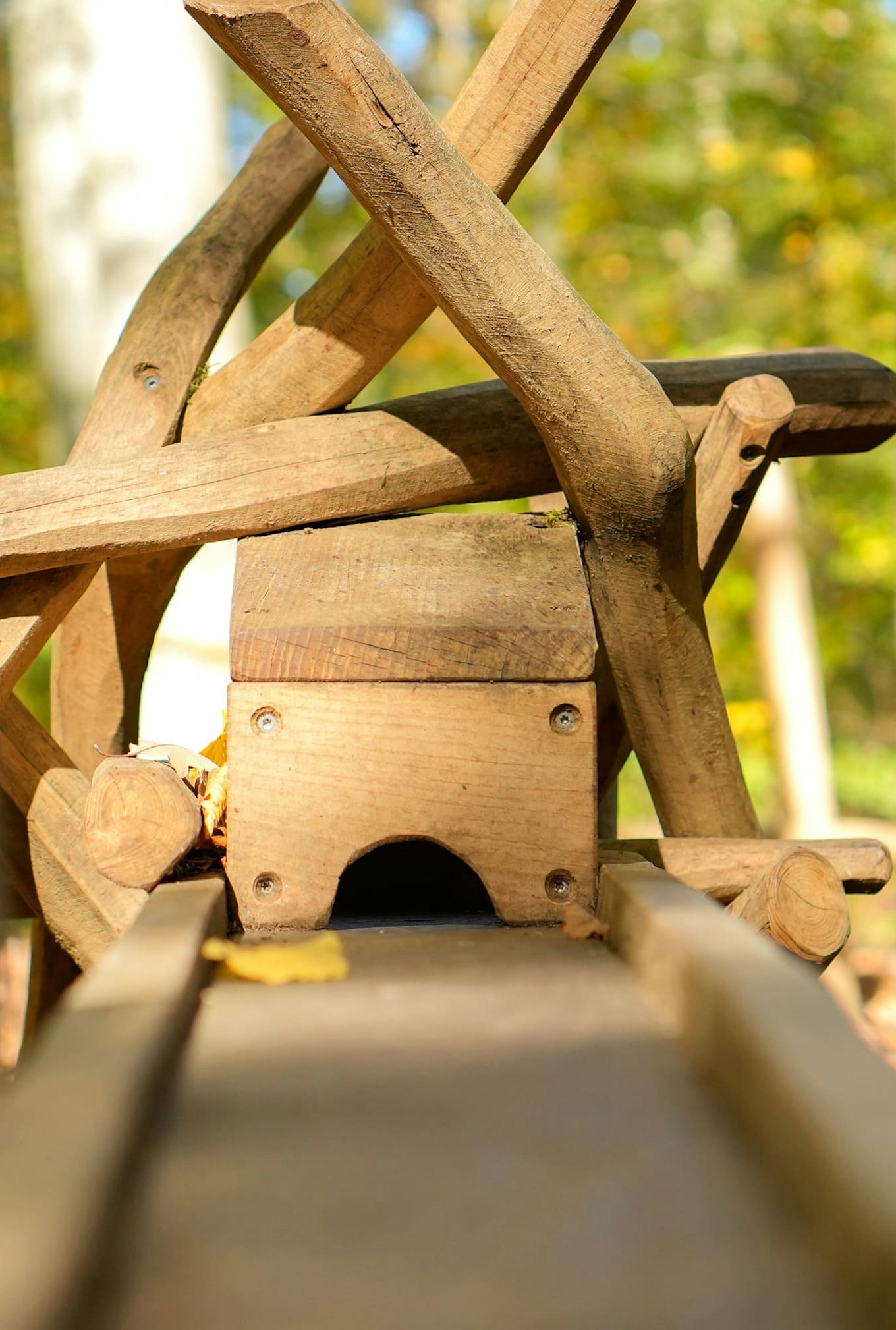 Nahaufnahme einer Holzkonstruktion mit Baumstämmen und Balken, die einem Spielplatz oder einer Skulptur in einer Waldlandschaft ähnelt. Sonnenlicht fällt durch die Bäume im Hintergrund.
