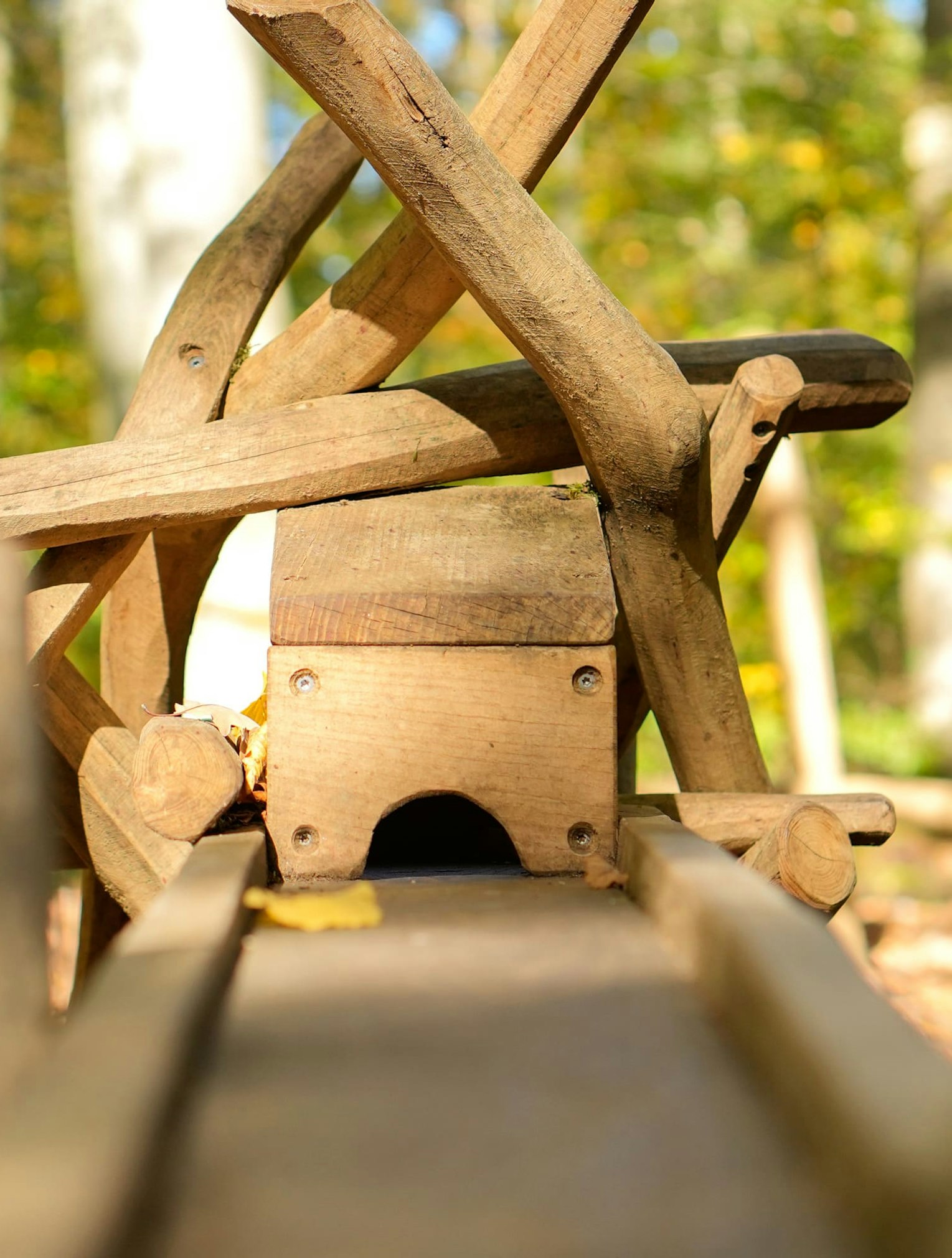 Nahaufnahme einer Holzkonstruktion mit Baumstämmen und Balken, die einem Spielplatz oder einer Skulptur in einer Waldlandschaft ähnelt. Sonnenlicht fällt durch die Bäume im Hintergrund.