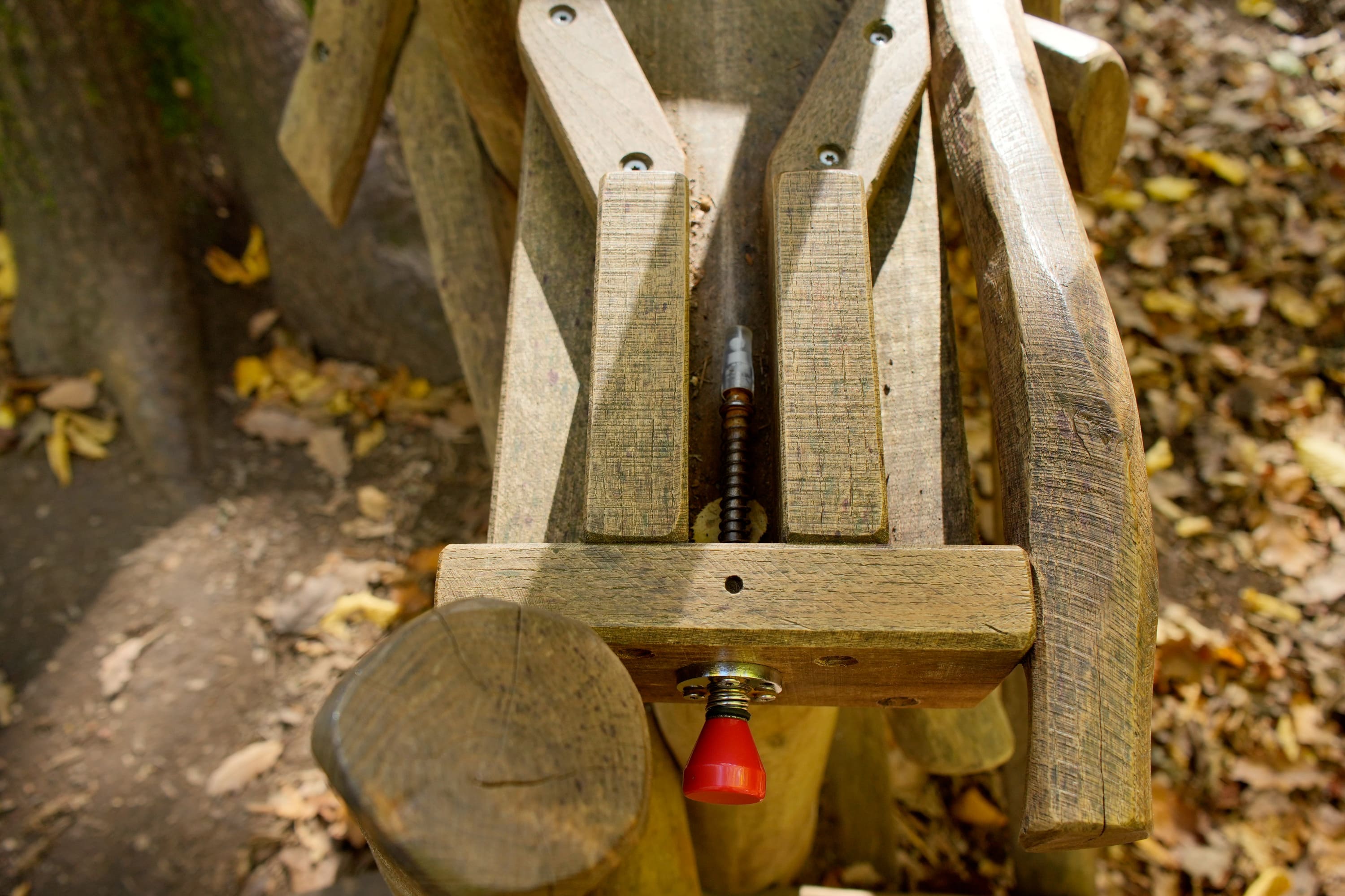 Murmelbahn mit einem Schrauben- und Scharniermechanismus aus Metall und einem roten Knopf. Umgeben von Laub auf dem Boden.