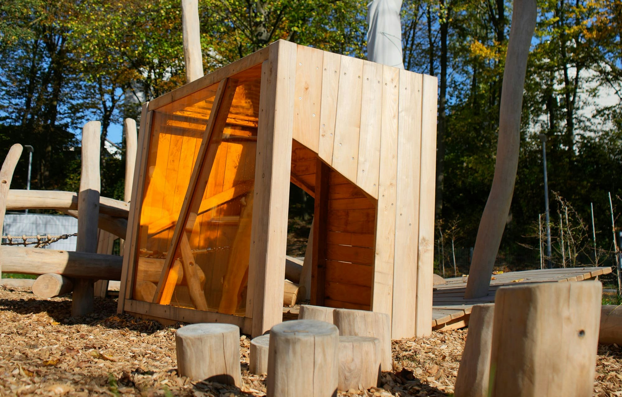 Eine hölzerne Spielplatzstruktur mit abgewinkelten Wänden und einer Rutsche steht umgeben von Holzspänen und Baumstümpfen vor einer Kulisse aus Bäumen mit Herbstlaub.
