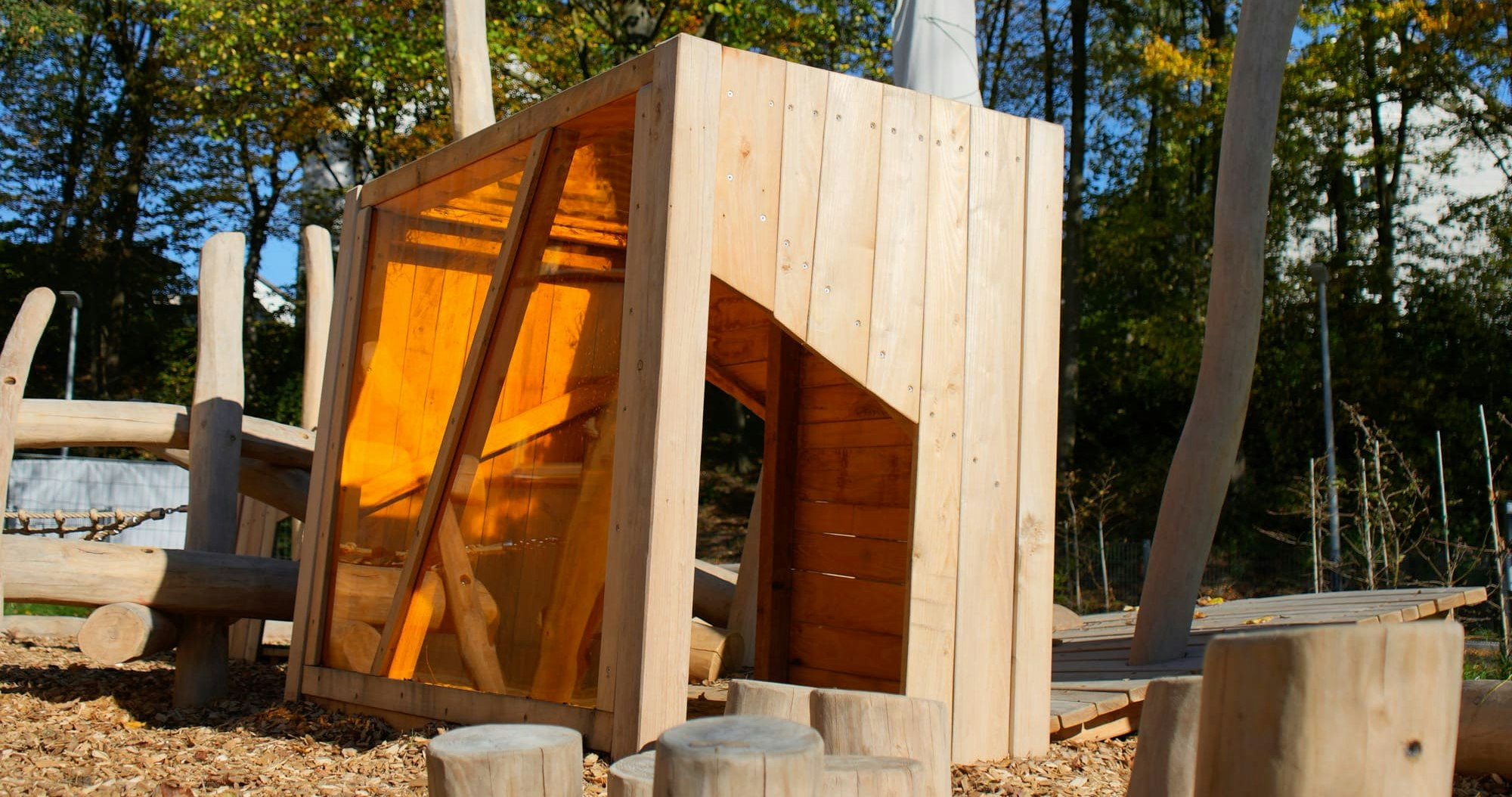 Eine hölzerne Spielplatzstruktur mit abgewinkelten Wänden und einer Rutsche steht umgeben von Holzspänen und Baumstümpfen vor einer Kulisse aus Bäumen mit Herbstlaub.