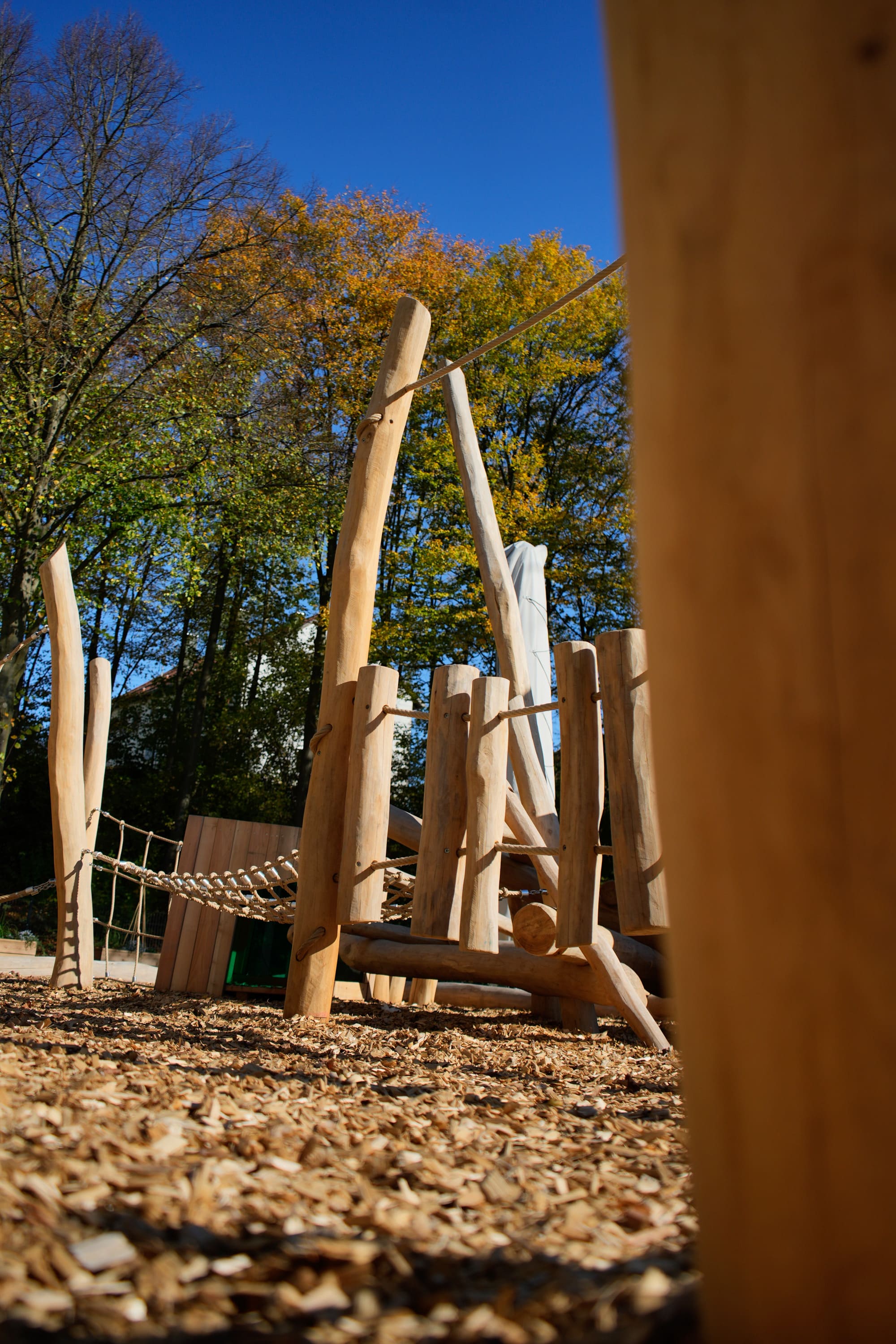 Hölzerne Spielplatzstruktur mit Kletterelementen und Seilbrücke, umgeben von Bäumen mit Herbstlaub unter einem klaren blauen Himmel.