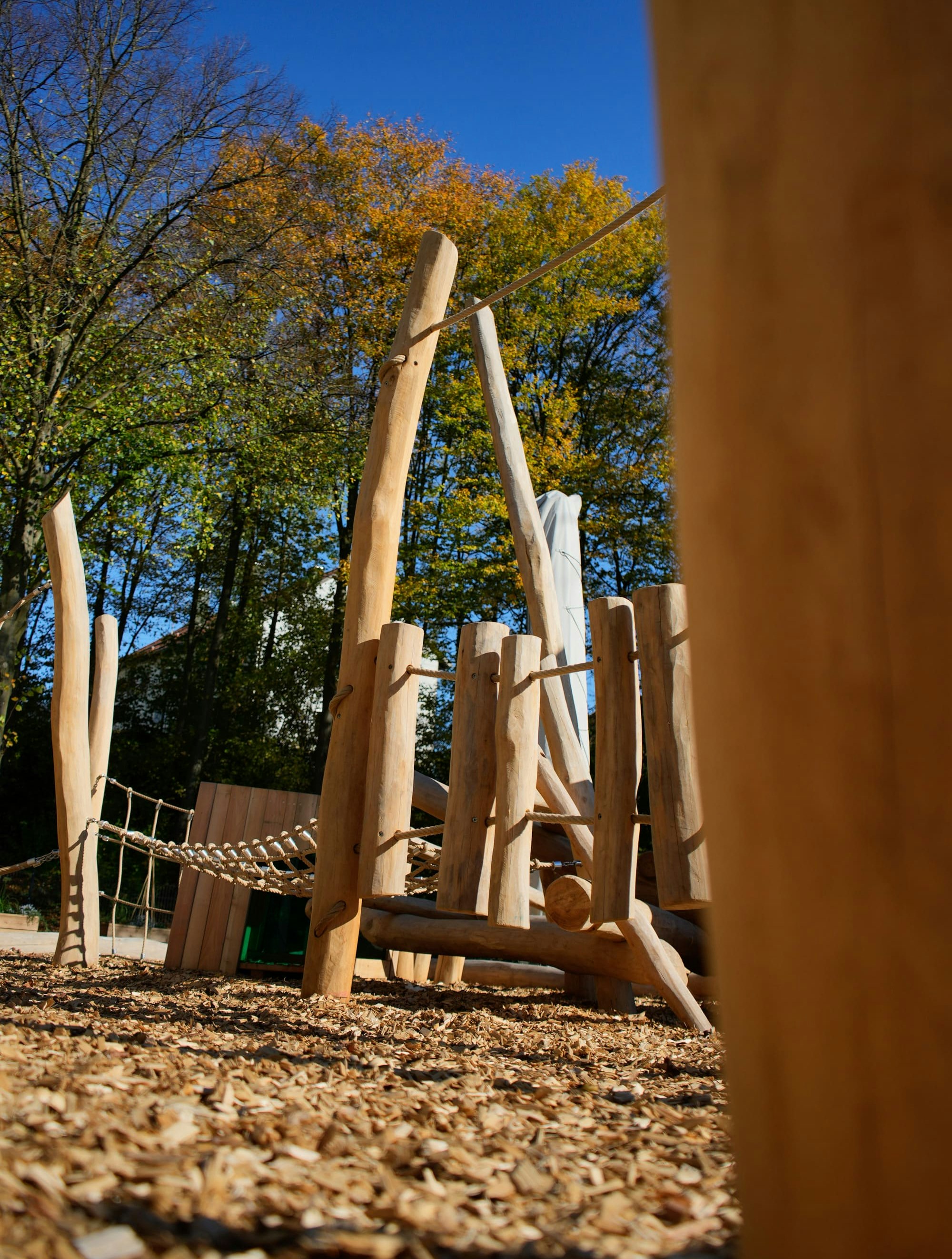 Hölzerne Spielplatzstruktur mit Kletterelementen und Seilbrücke, umgeben von Bäumen mit Herbstlaub unter einem klaren blauen Himmel.